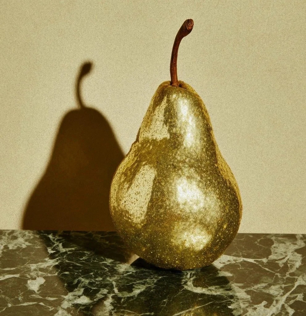A pear with a glittery or metallic surface standing on a marble surface, casting a distinct shadow on the wall behind it.