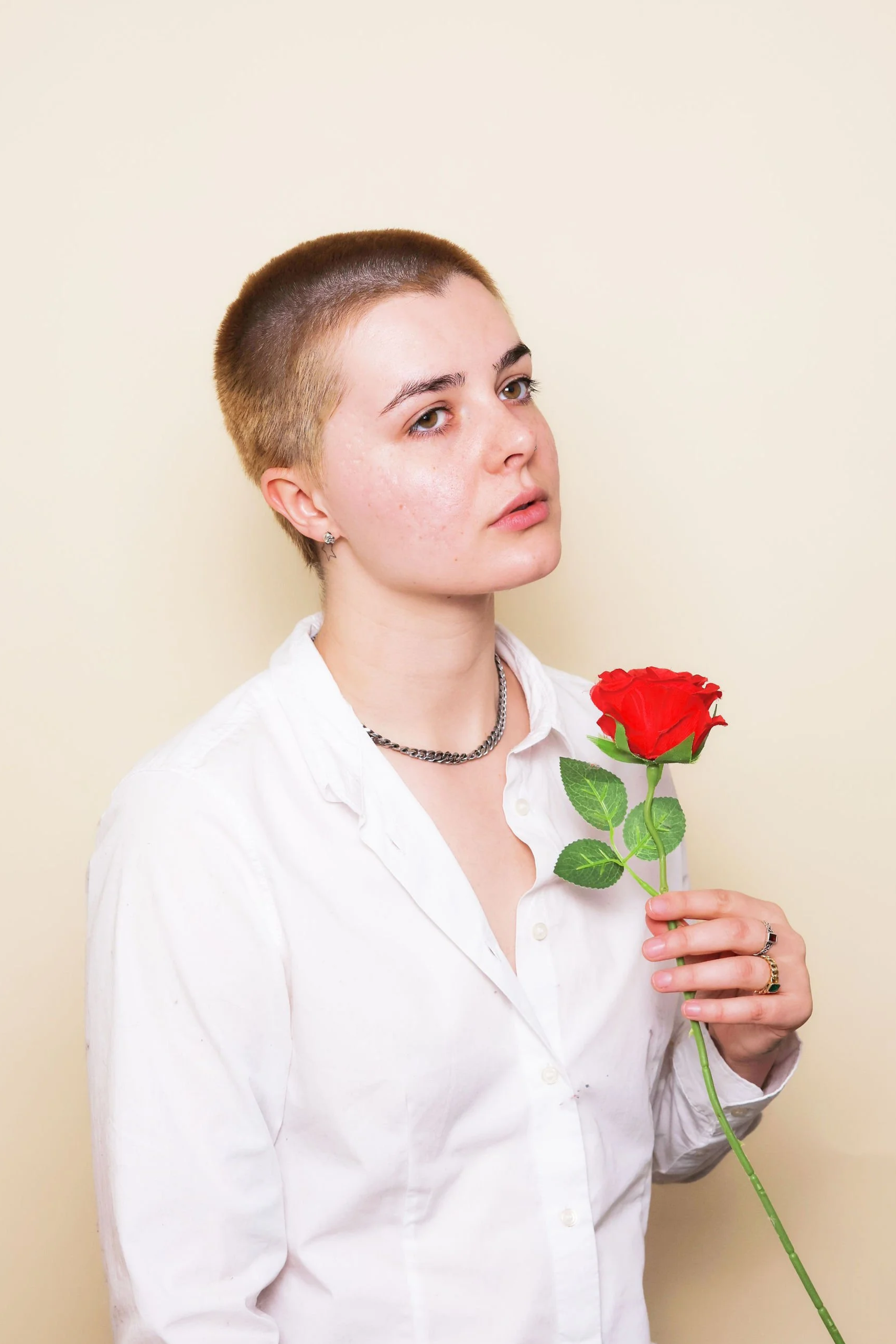 A young person with short hair holding a red rose, wearing a white shirt, a silver chain necklace, and rings, standing against a beige background.