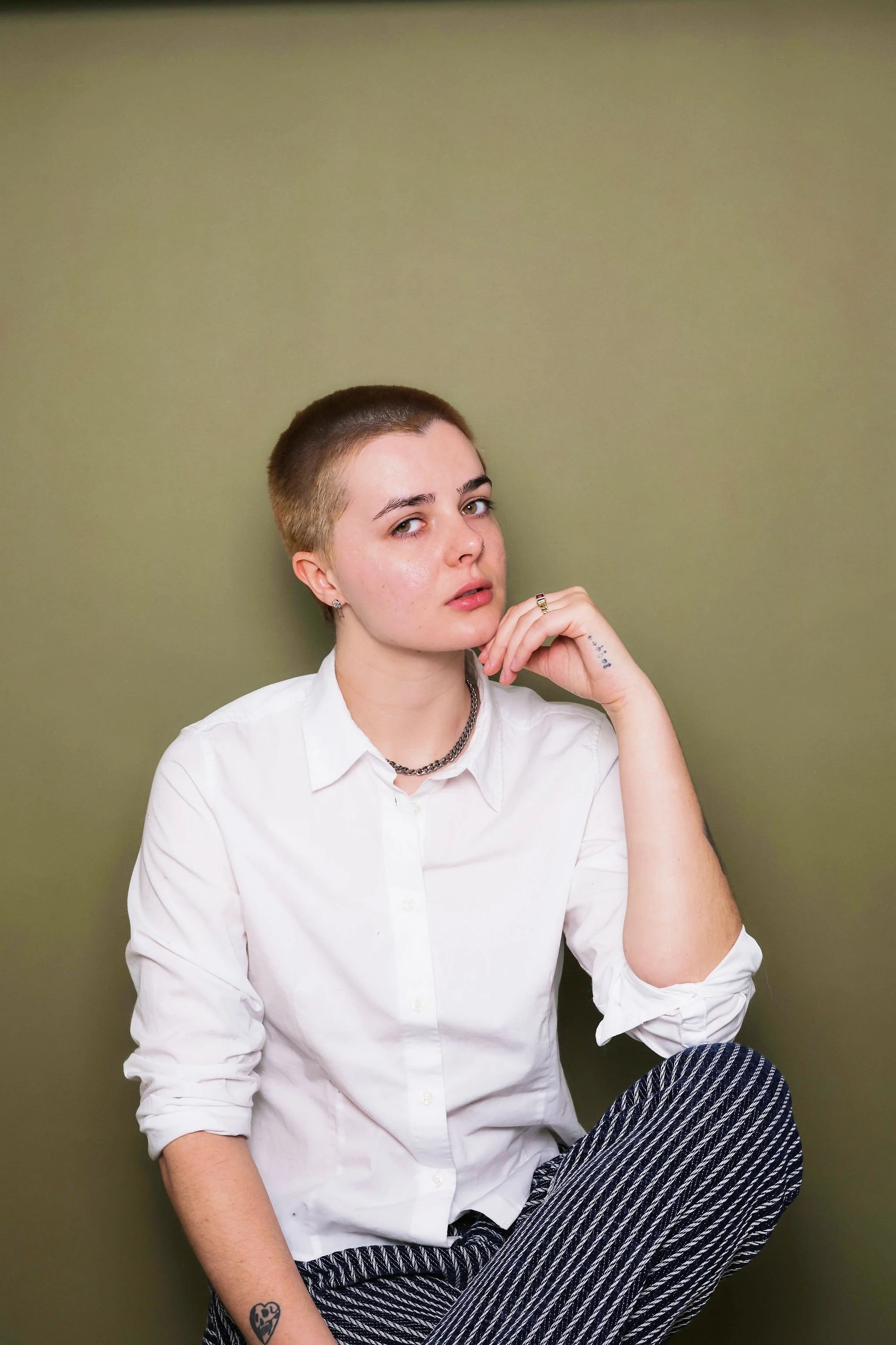 Young person with short hair, wearing a white button-up shirt and patterned pants, sitting against a plain green wall, looking at the camera with a contemplative expression.