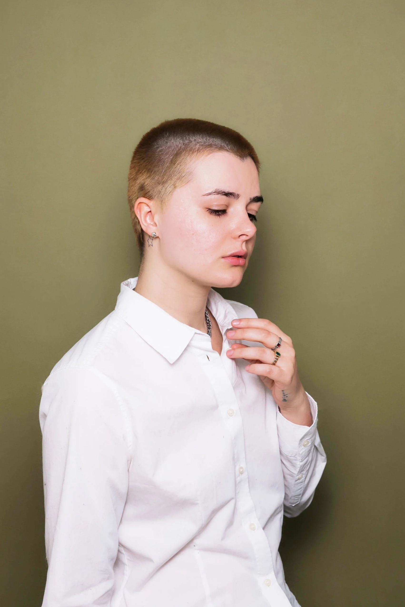 Young woman with a short haircut wearing a white shirt, looking down with a neutral expression against a plain background.