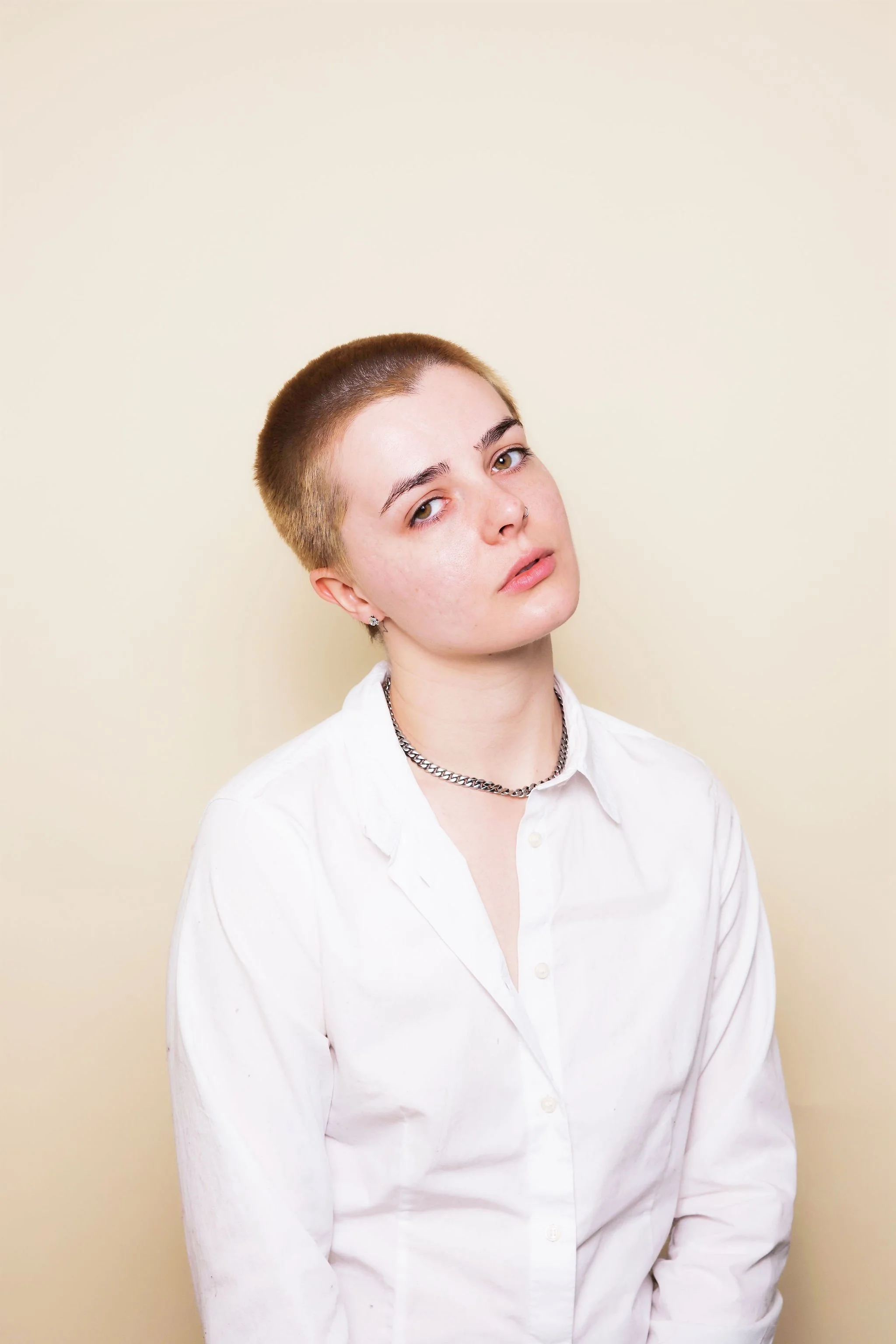 Young woman with a shaved head, wearing a white shirt, silver chain necklace, and earrings, posing against a beige background.