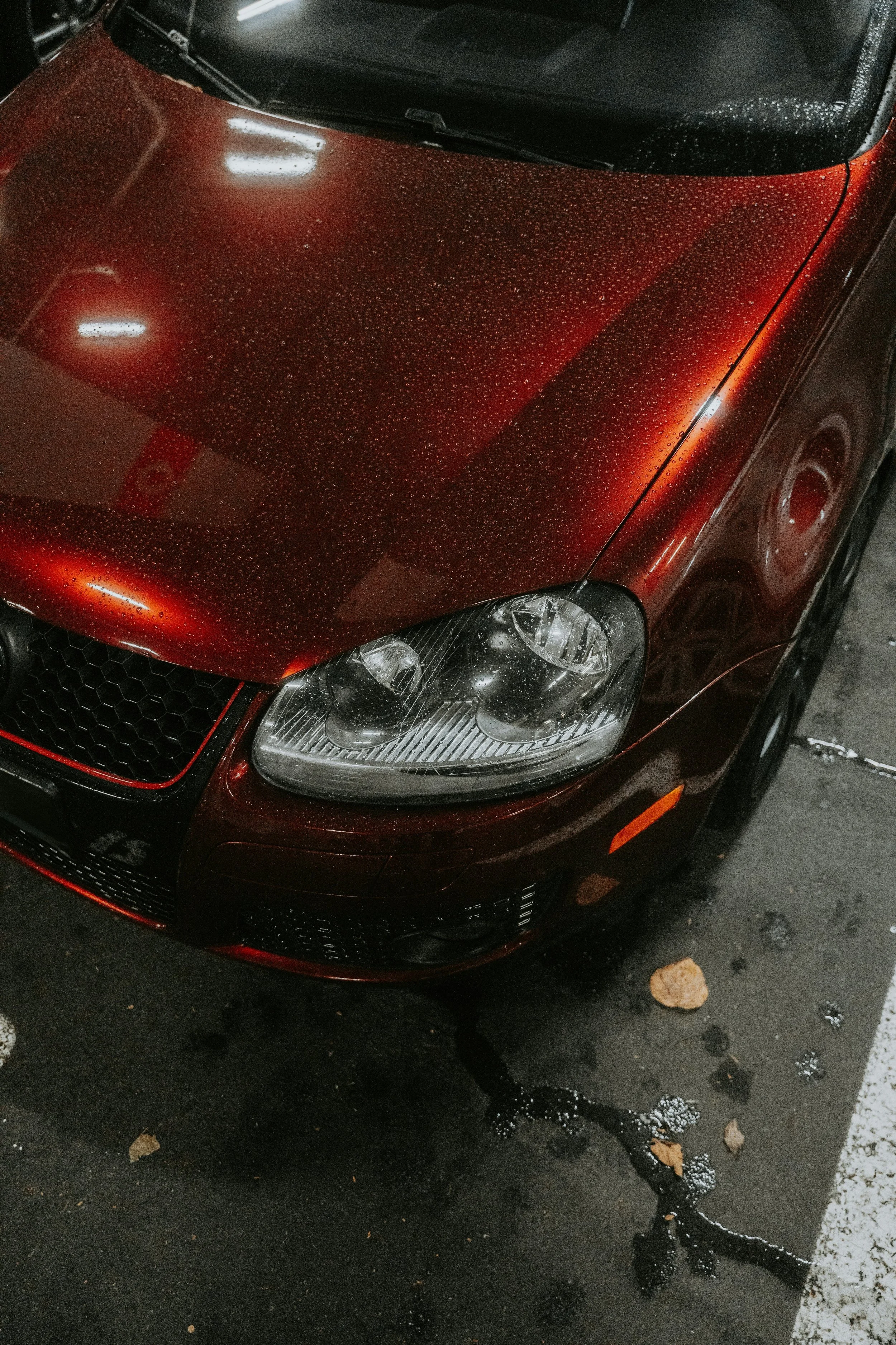 A close-up of a red car with raindrops on the hood and headlight, parked in a parking lot with some water and rocks on the ground.