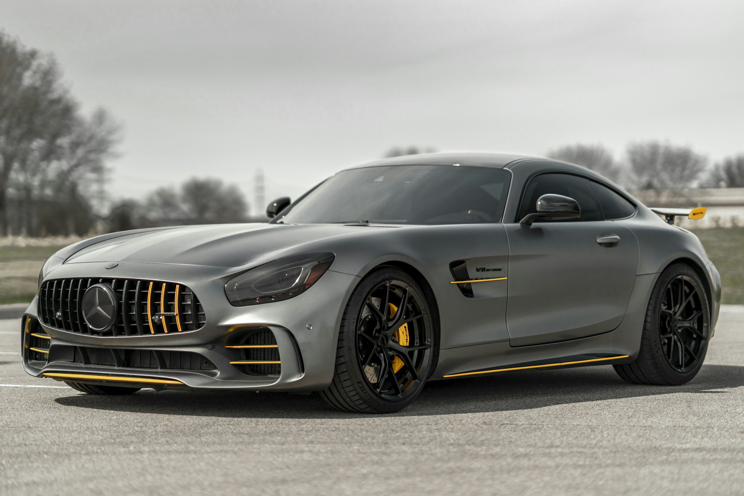 A silver Mercedes-Benz sports car with black wheels and yellow accents, parked outdoors on a paved surface, with trees and an overcast sky in the background.