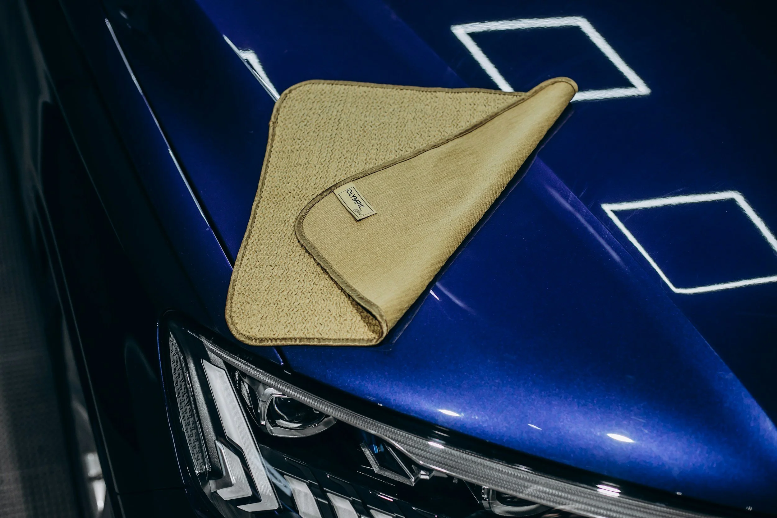 A gold-colored car cleaning or polishing cloth with a label that says 'OLYMPIC' rests on the hood of a shiny blue car.