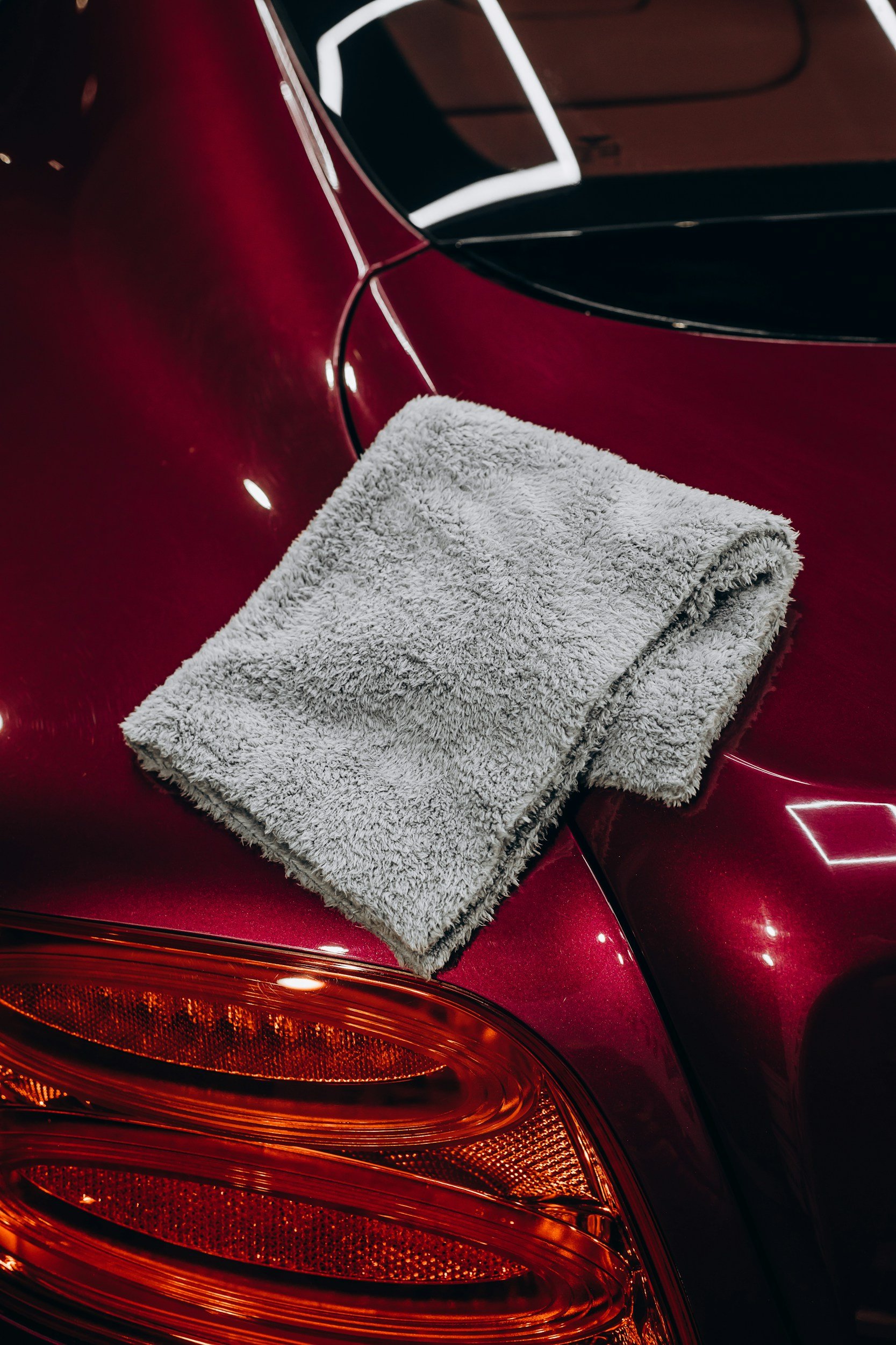 A gray towel laid on the trunk of a shiny red car, near the taillight and rear window.