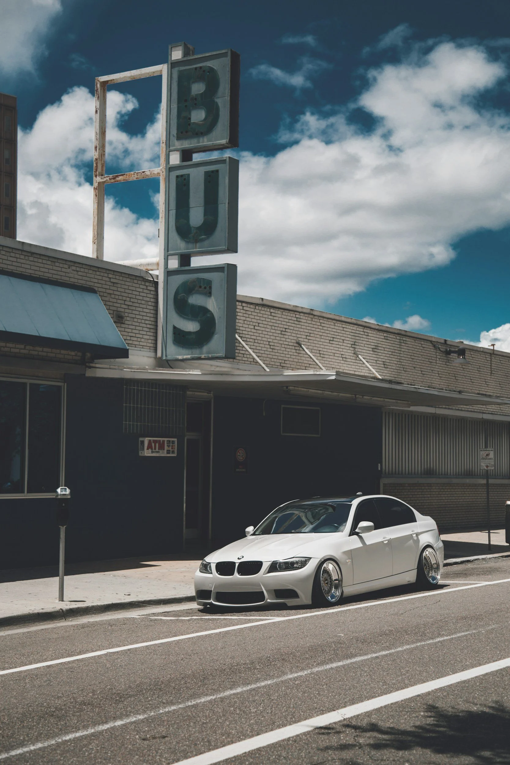 A silver lowered BMW coupe parked in front of a building with a large vintage sign that reads 'BULS' against a blue sky with clouds.