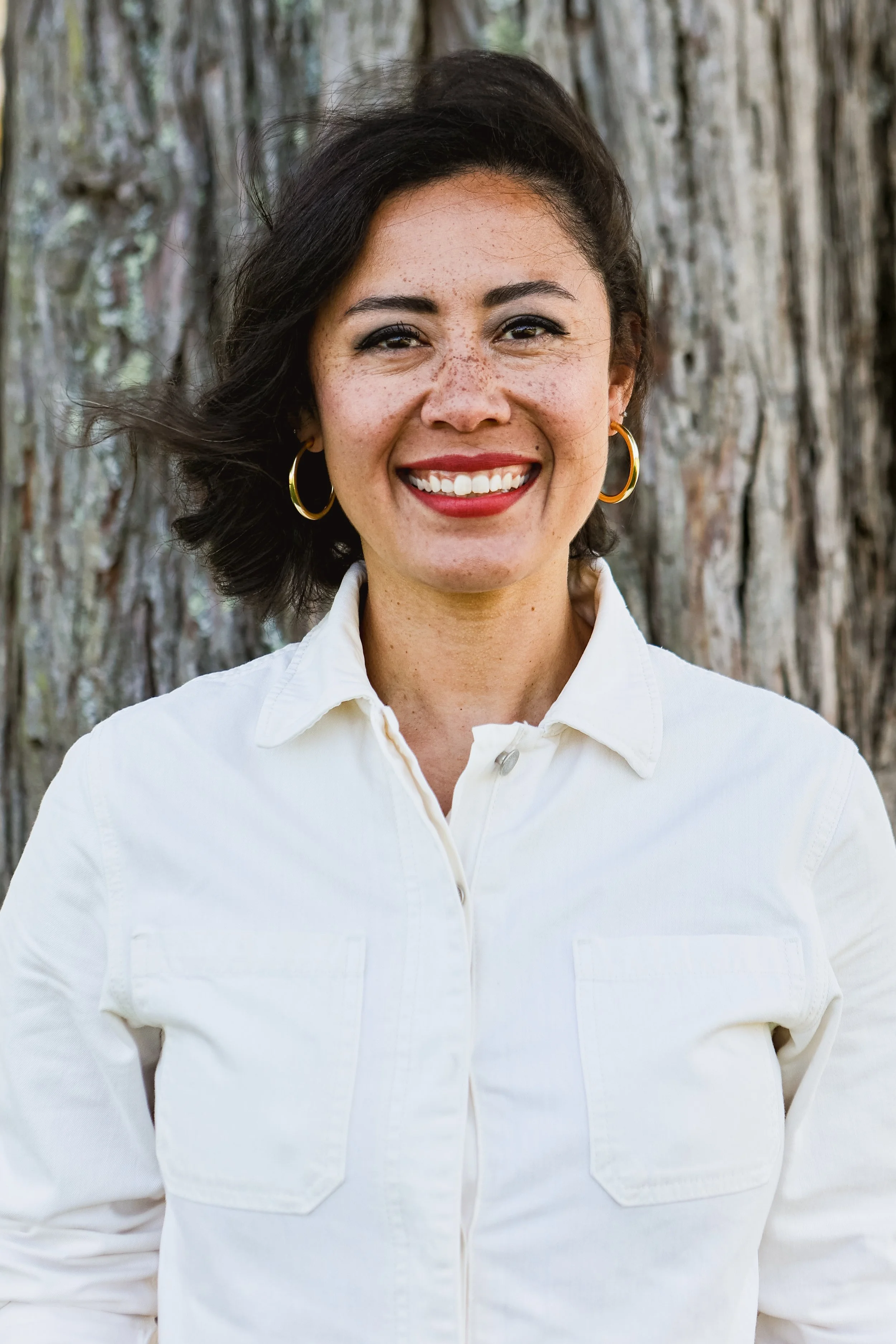A woman smiling outdoors with a large tree trunk in the background. She has shoulder-length dark hair, is wearing gold hoop earrings, bright red lipstick, and a white button-up shirt.
