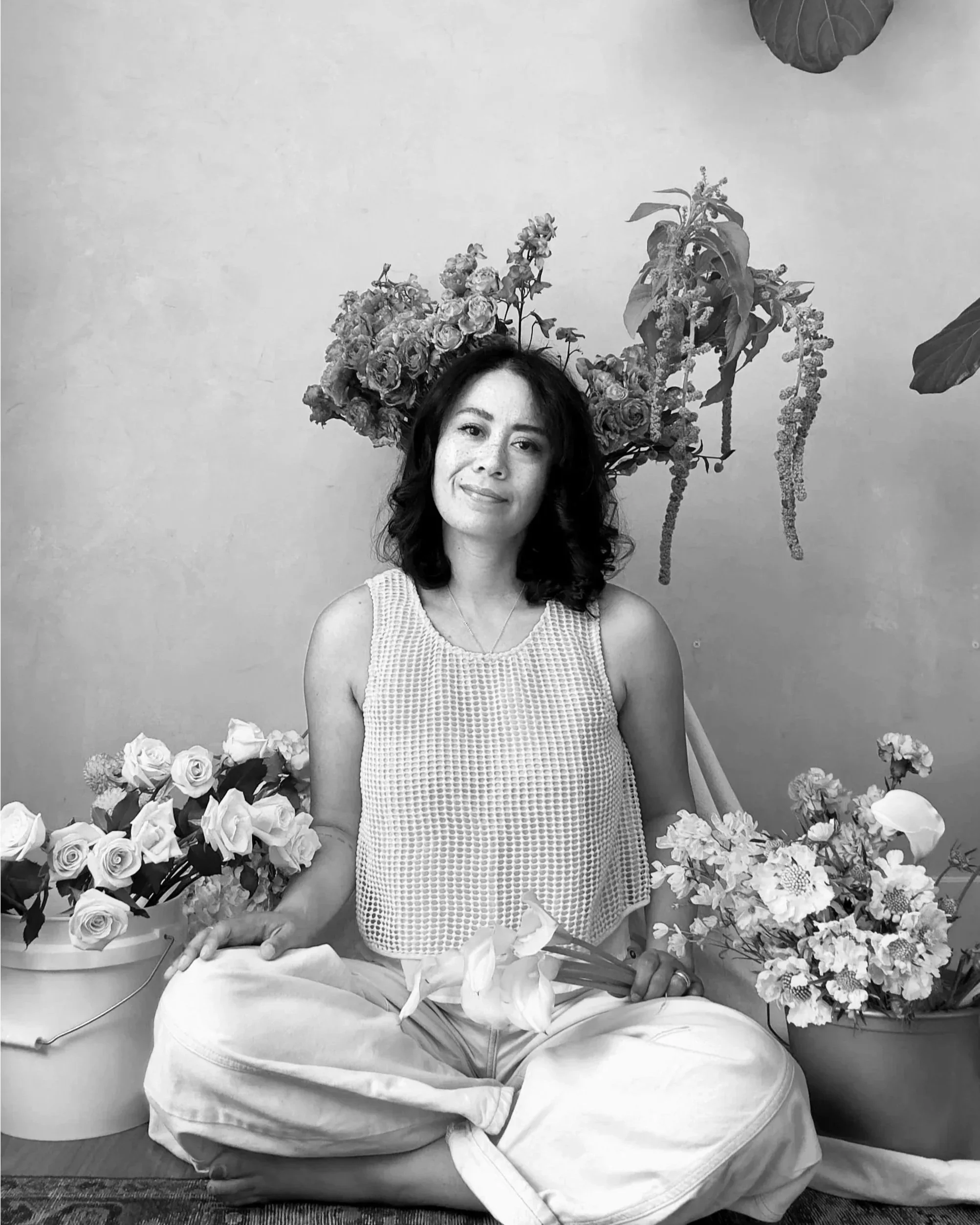 Lani sitting cross-legged with flowers around her, in front of a plain wall with hanging plants and flowers behind her.