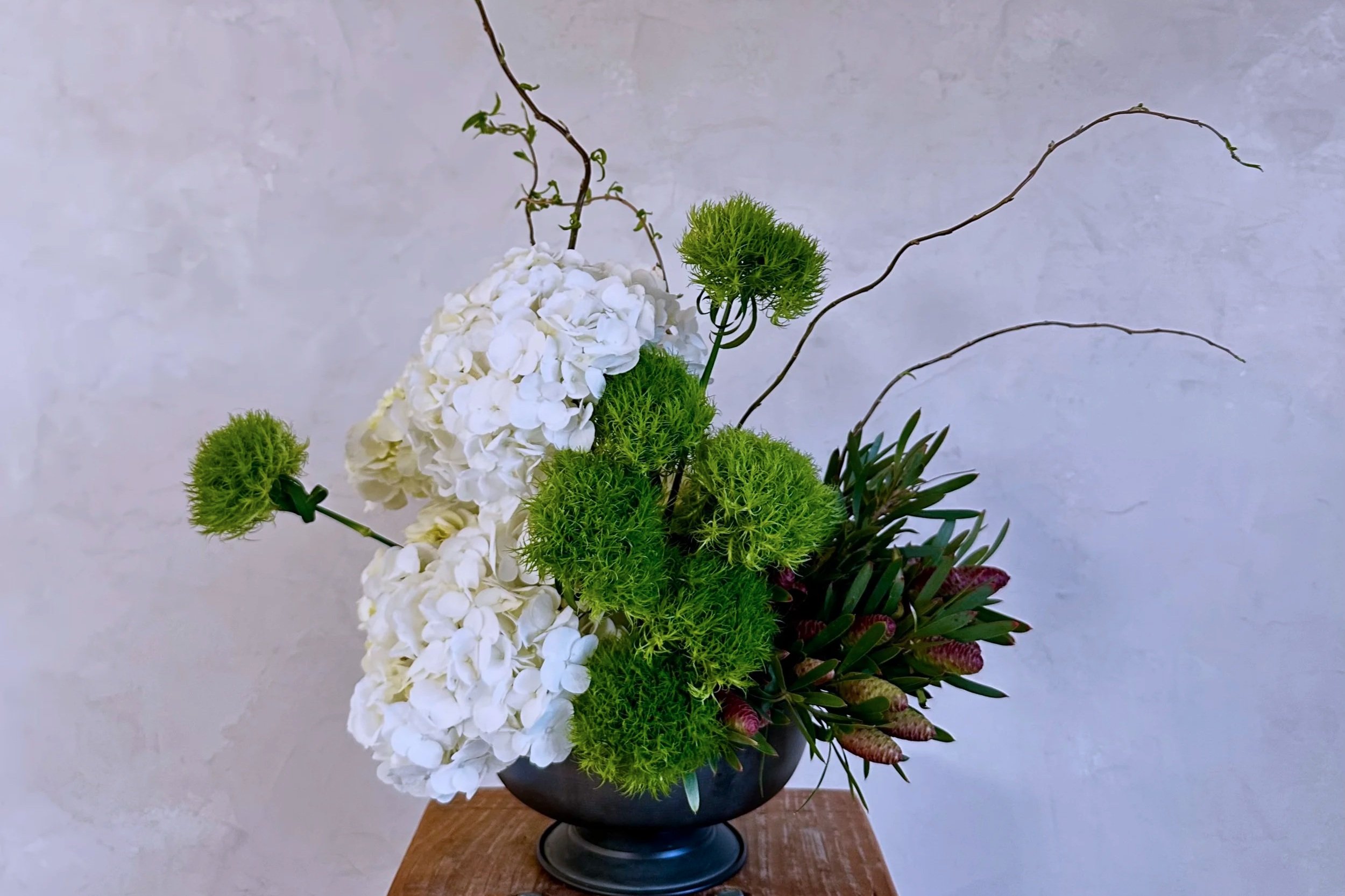 A floral arrangement in a black vase containing white hydrangeas, green allium flowers, and other greenery, set against a plain, light-colored wall background.