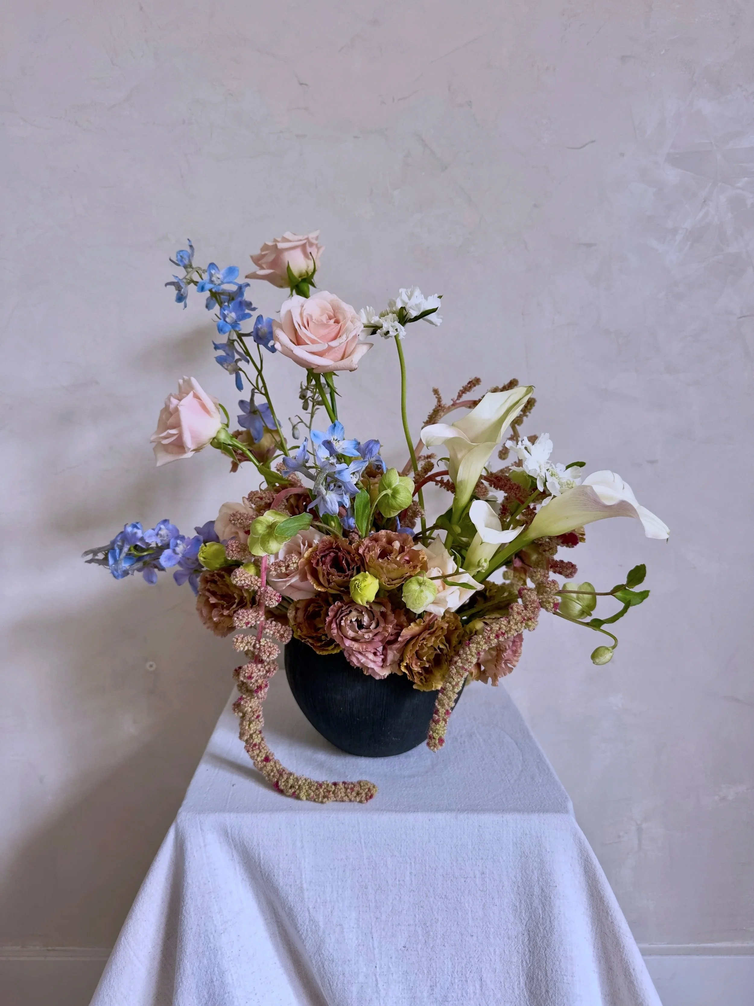 A floral arrangement with pink roses, white calla lilies, blue delphiniums, and other flowers in a black vase on a white table.