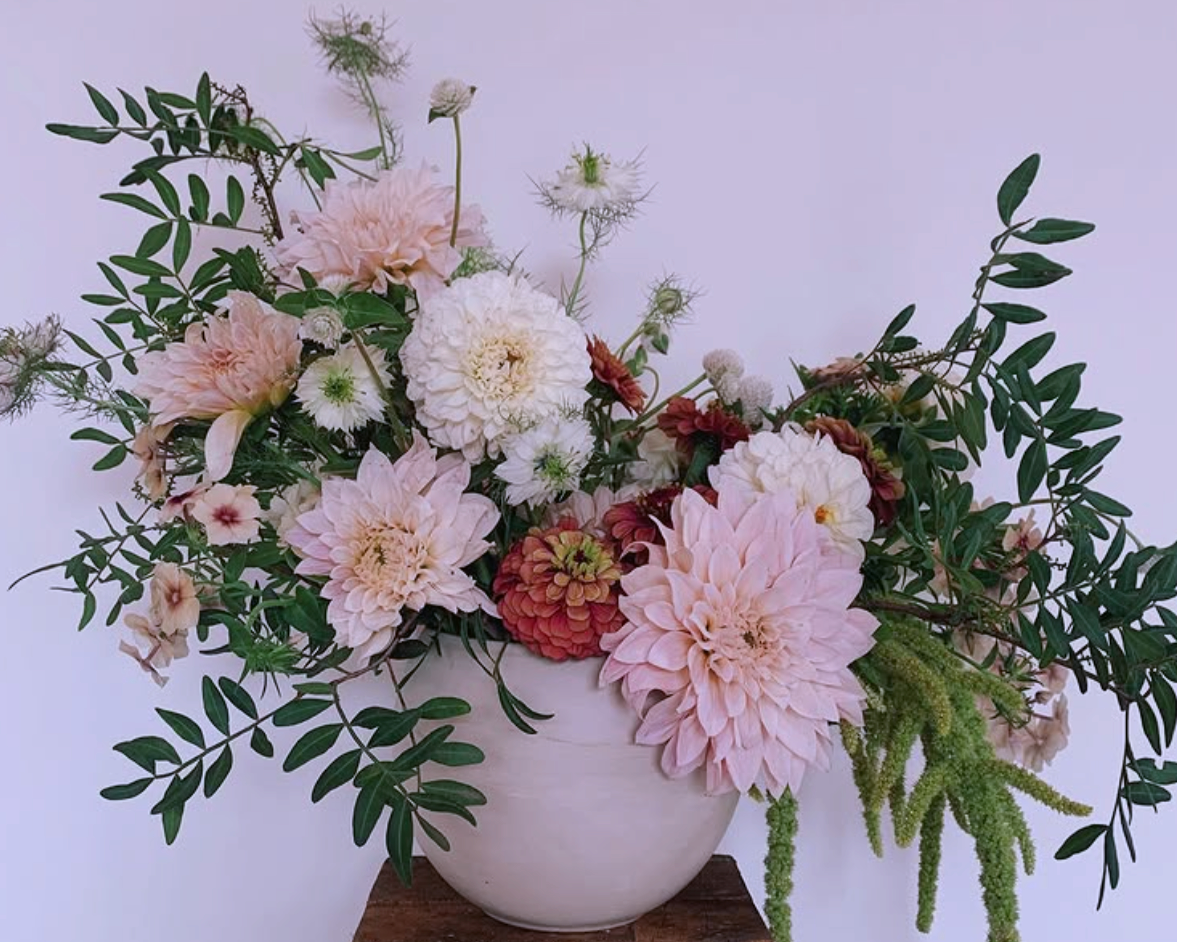 A white ceramic vase filled with pink, white, and red flowers, with green foliage, placed on a wooden surface against a plain white background.