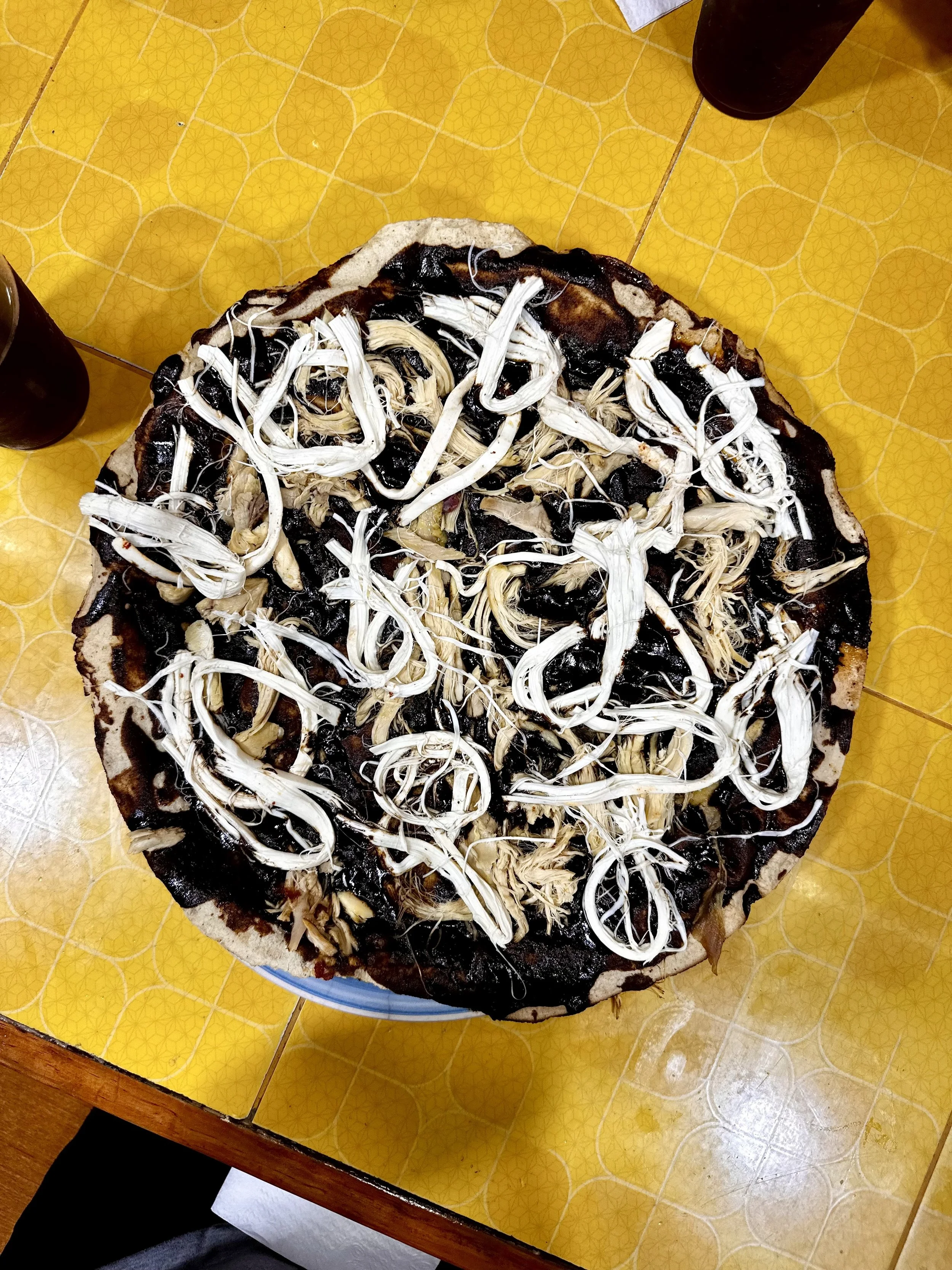 A pizza with a dark base, topped with shredded white cheese and black ingredients, possibly mushrooms or olives, on a yellow tile table.