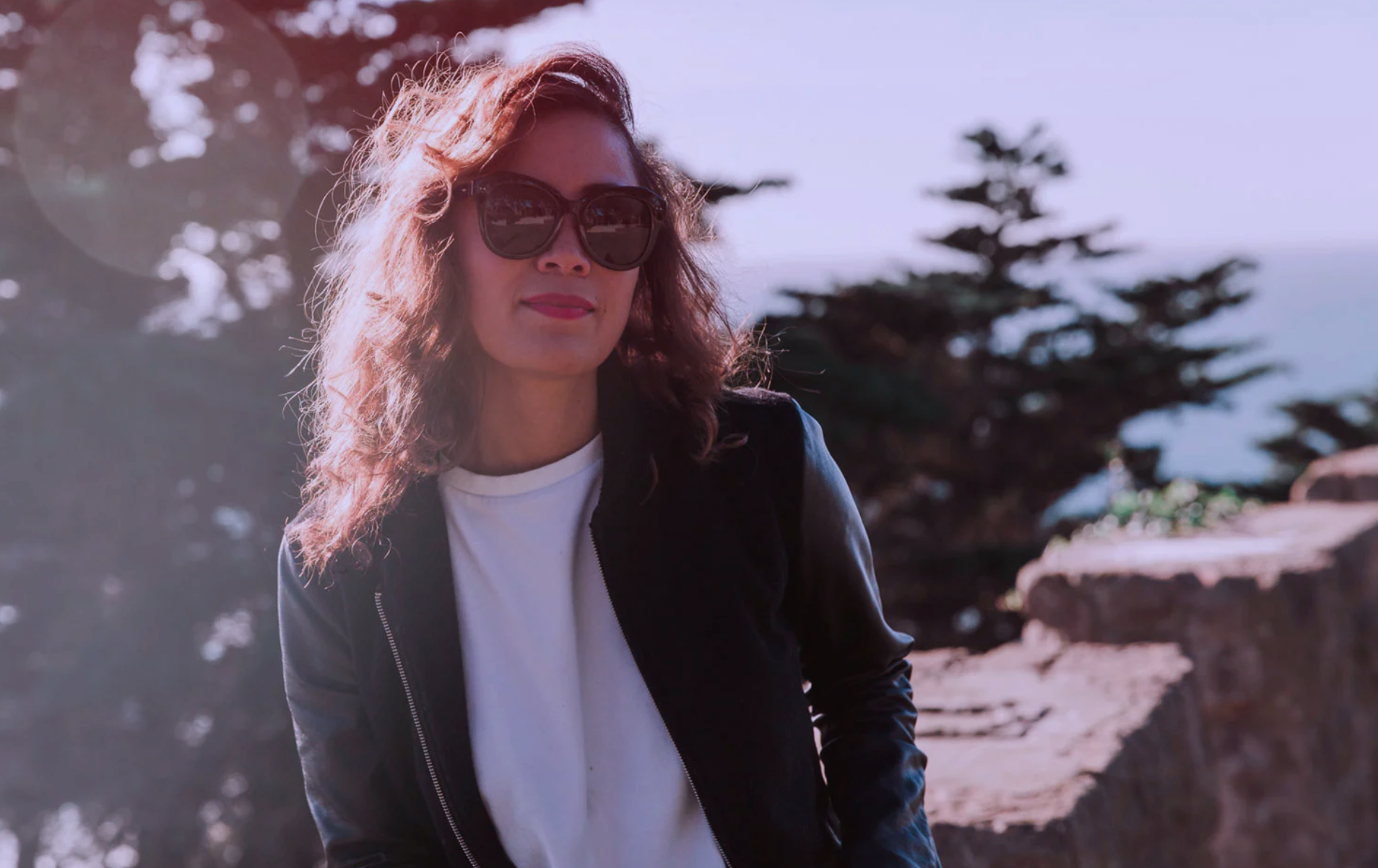 A woman with curly hair wearing sunglasses and a black jacket stands outdoors near a stone wall with trees in the background during the daytime.