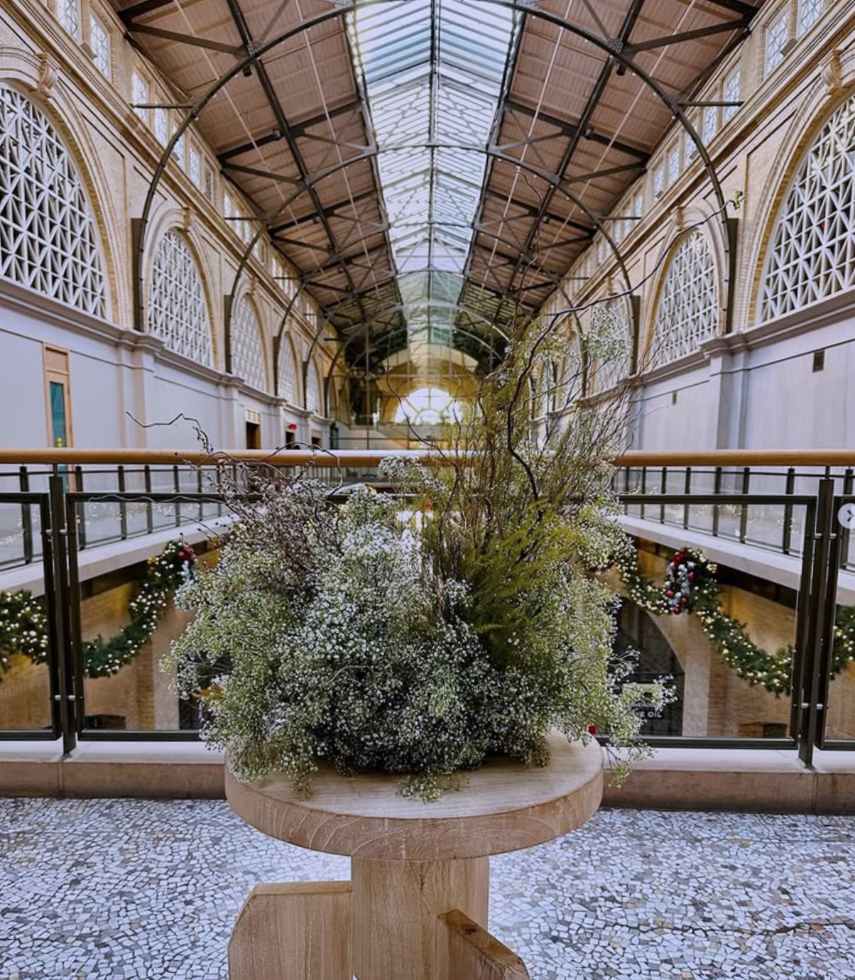 A large indoor space with a high, arched glass ceiling, ornate architectural details, and holiday decorations. In the foreground, a round wooden table holds a large floral arrangement of white flowers and greenery. The background includes a balcony with holiday garlands and wreaths.