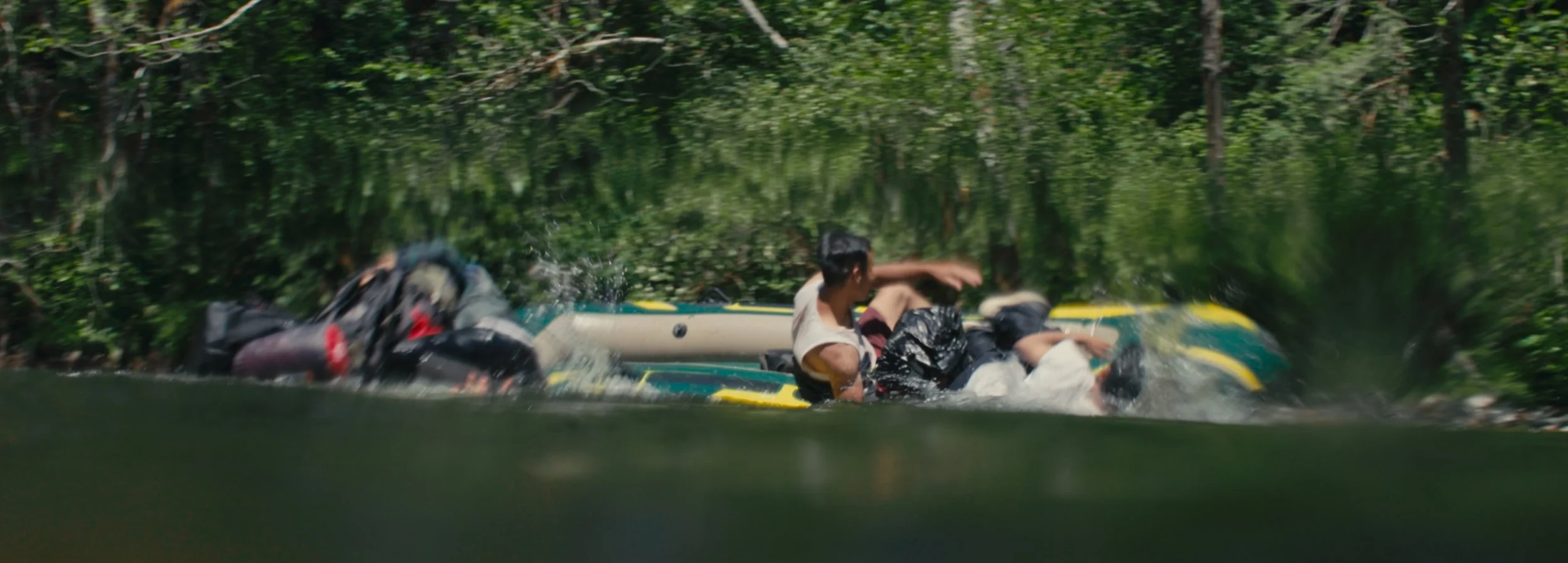 Two people falling into the water from an inflatable boat in a lush green river environment.