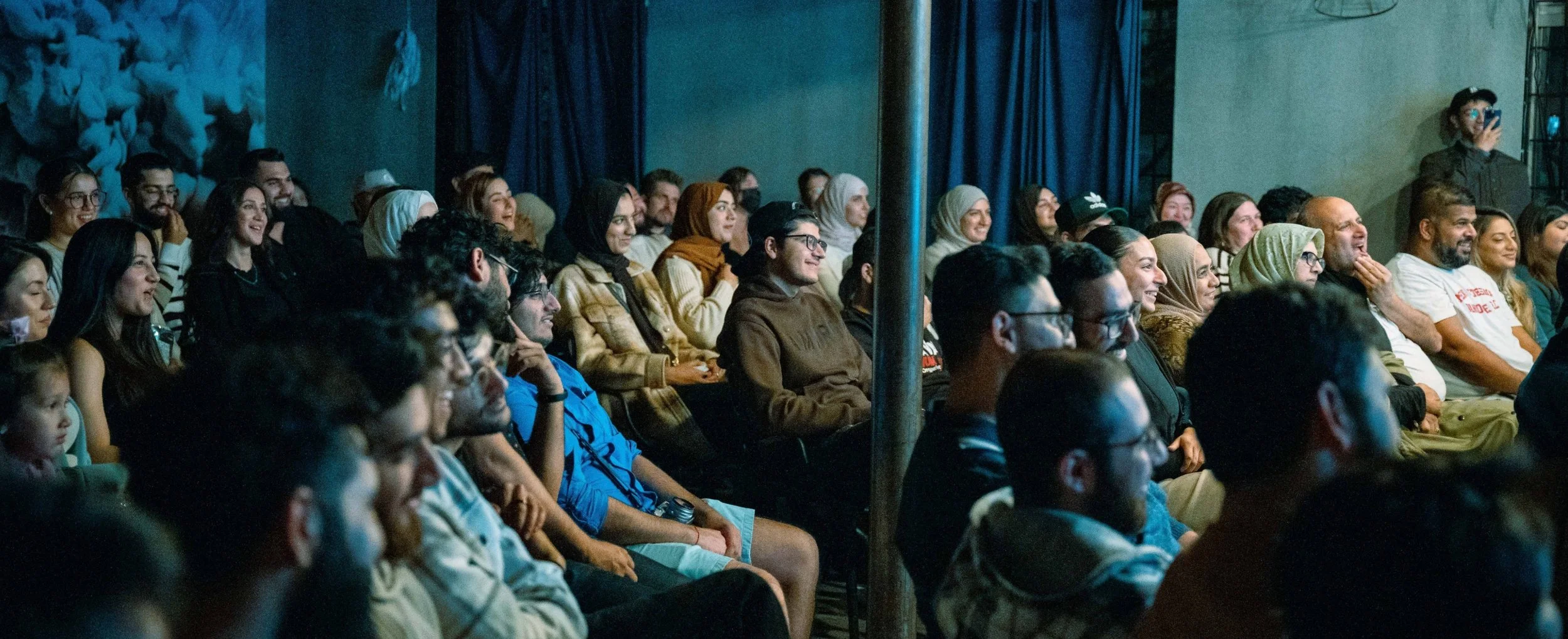 Audience seated in a dimly lit auditorium, watching a performance or presentation, with diverse individuals smiling and engaged.
