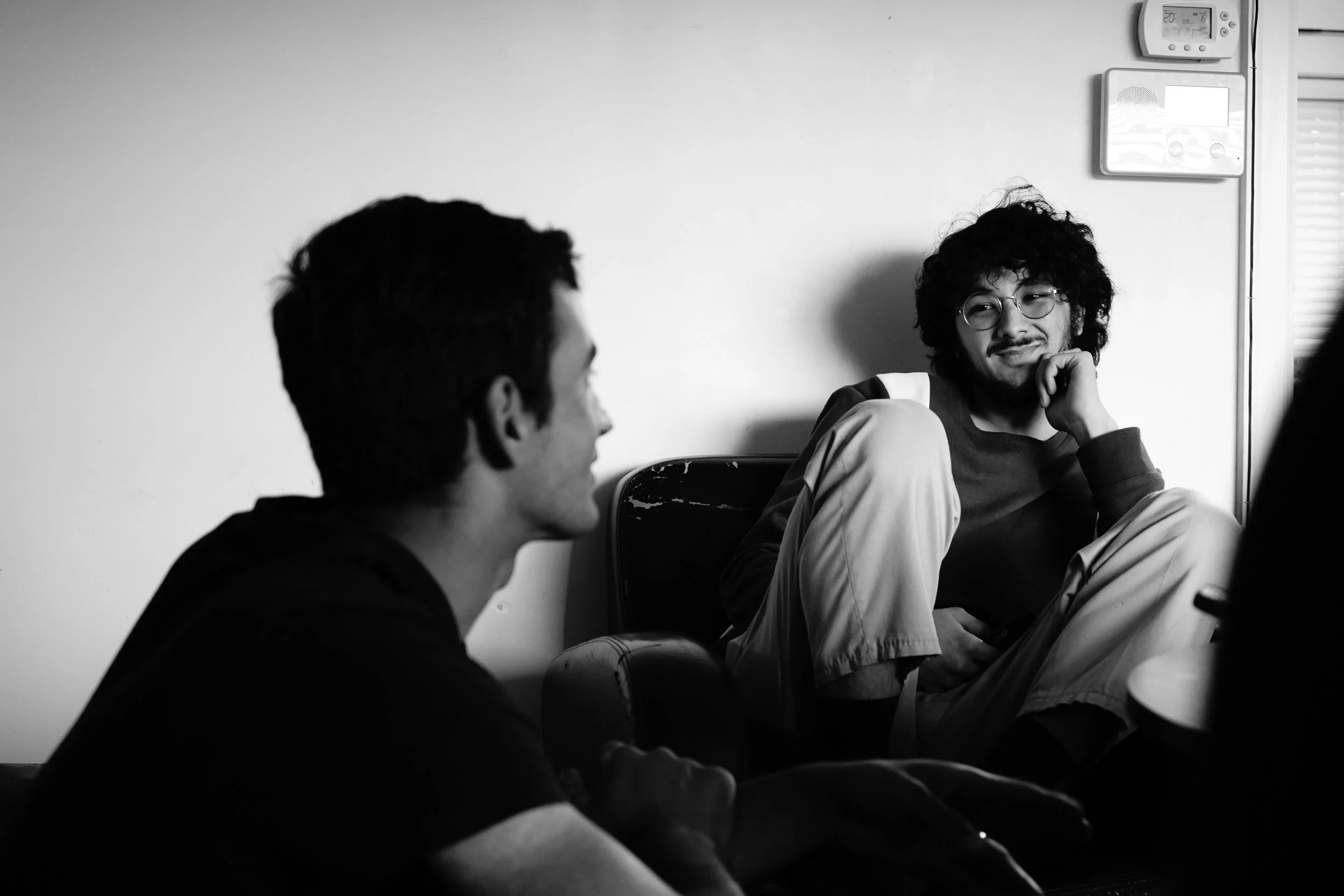 Two young men sitting and talking in a room, one with curly hair and glasses, the other with straight hair, both casually dressed.