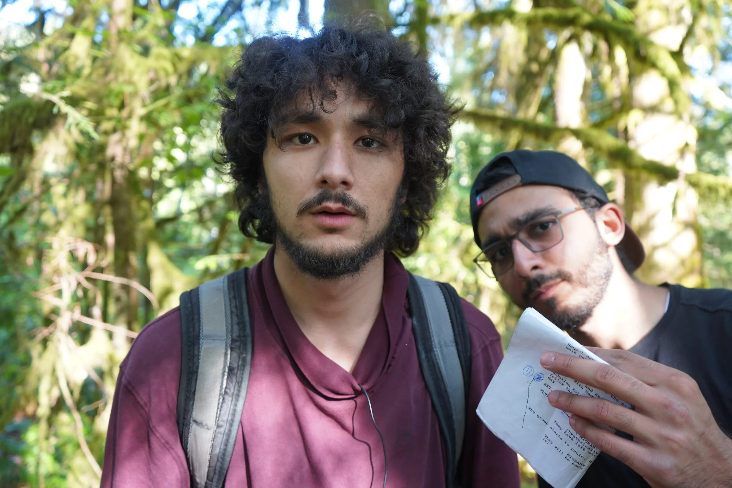 Two young men outdoors in a forest, one with curly hair and a backpack, the other with glasses and a cap, holding a crumpled sheet of paper and looking at the camera.