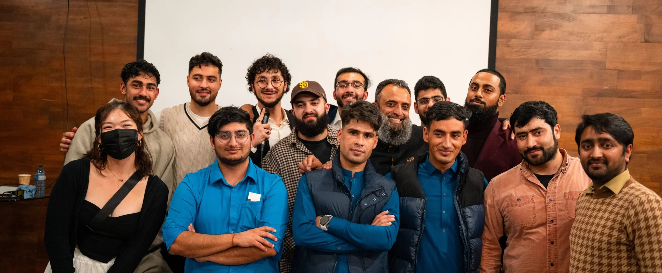 A diverse group of fifteen people posing for a photo indoors against a wooden wall and a white projection screen. Some individuals are smiling, one woman is wearing a black face mask, and one person is holding a small microphone.