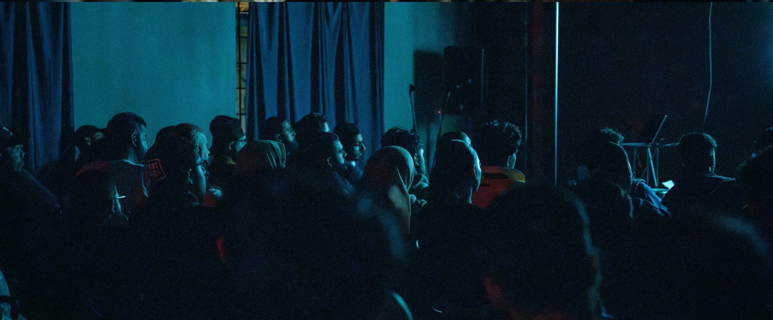 Audience watching a performance or presentation in a dark theater or auditorium, illuminated by a blue light.