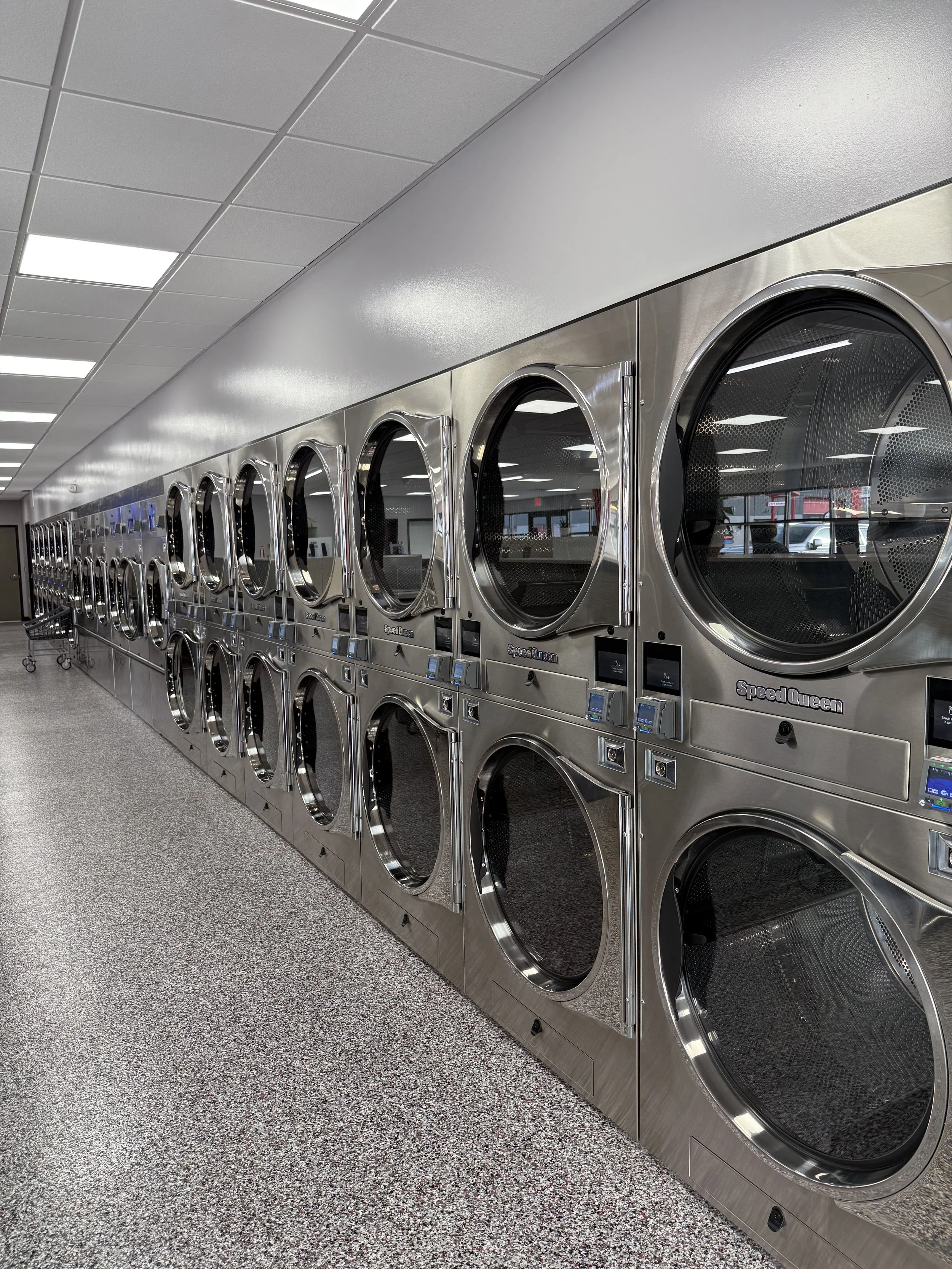 A row of large stainless steel commercial washing machines in a laundromat.