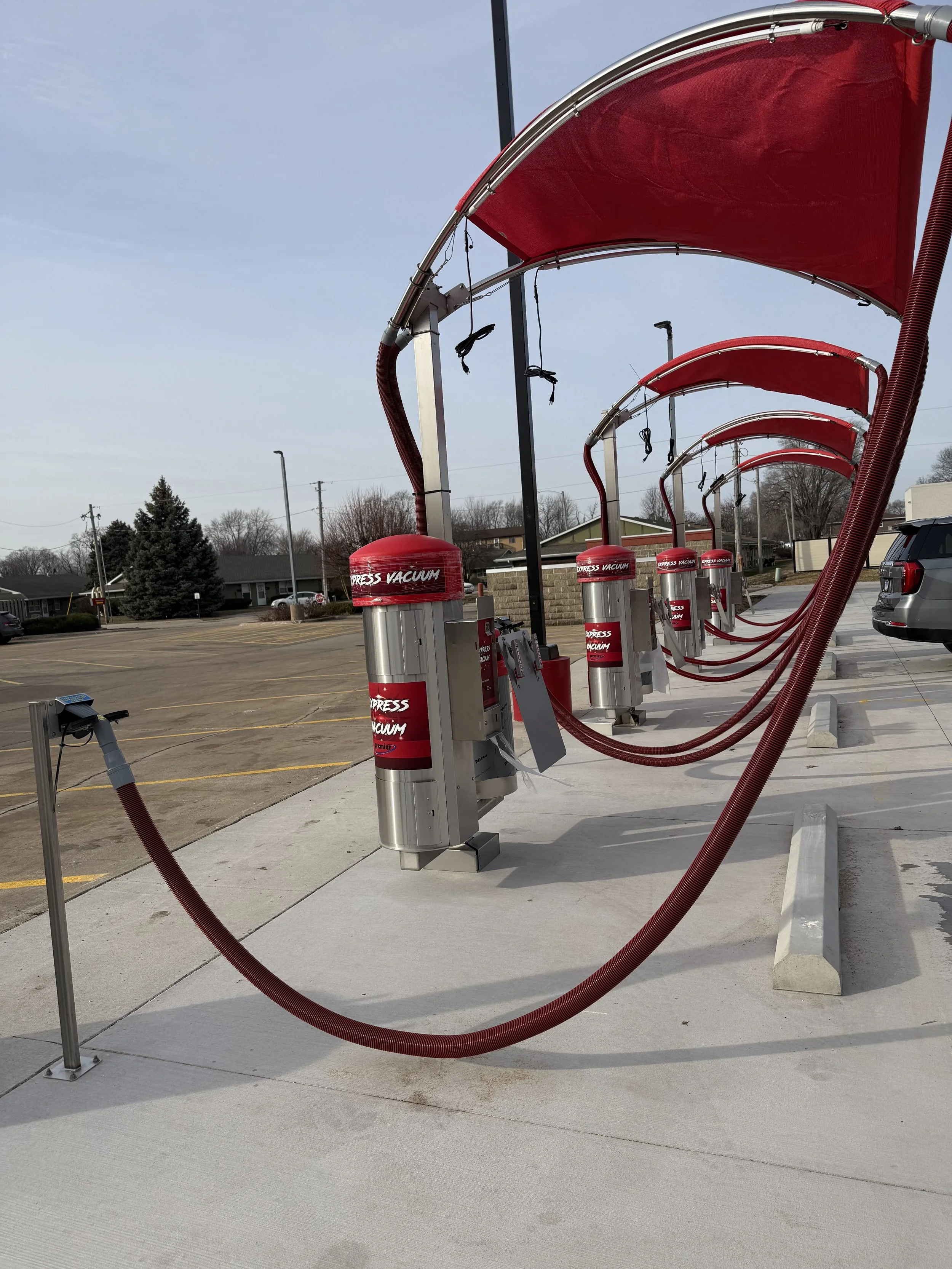 Self-service car wash station with multiple vacuum cleaners, red canopies, and hoses in a parking lot on a cloudy day.