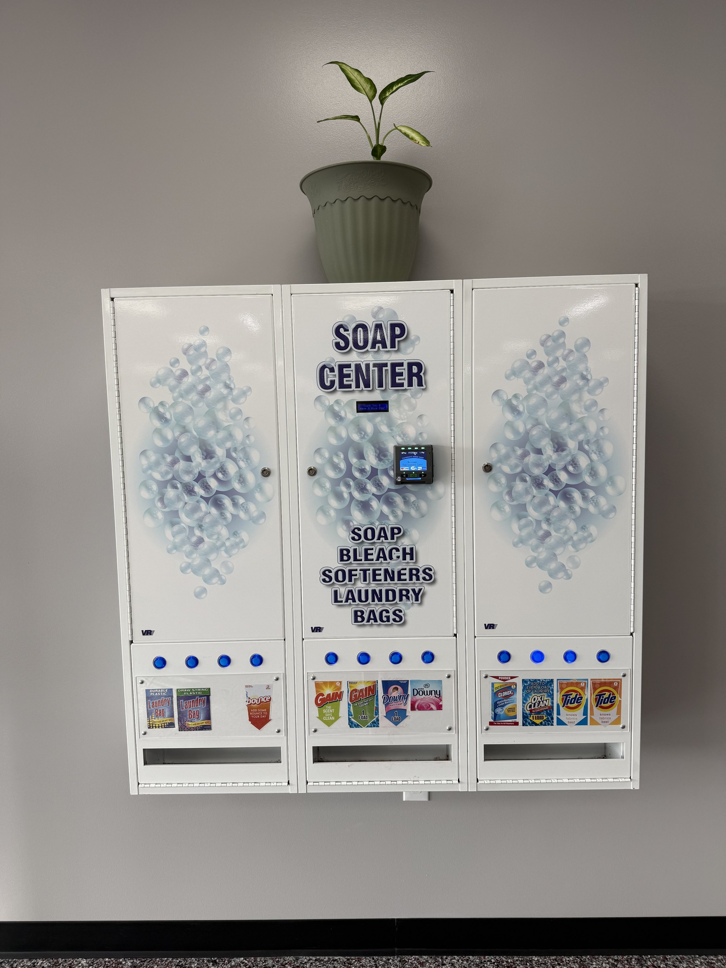 Wall-mounted soap vending machine labeled "Soap Center" with compartments for soap, bleach, softeners, laundry bags, and detergent packets beneath a potted plant on the wall.