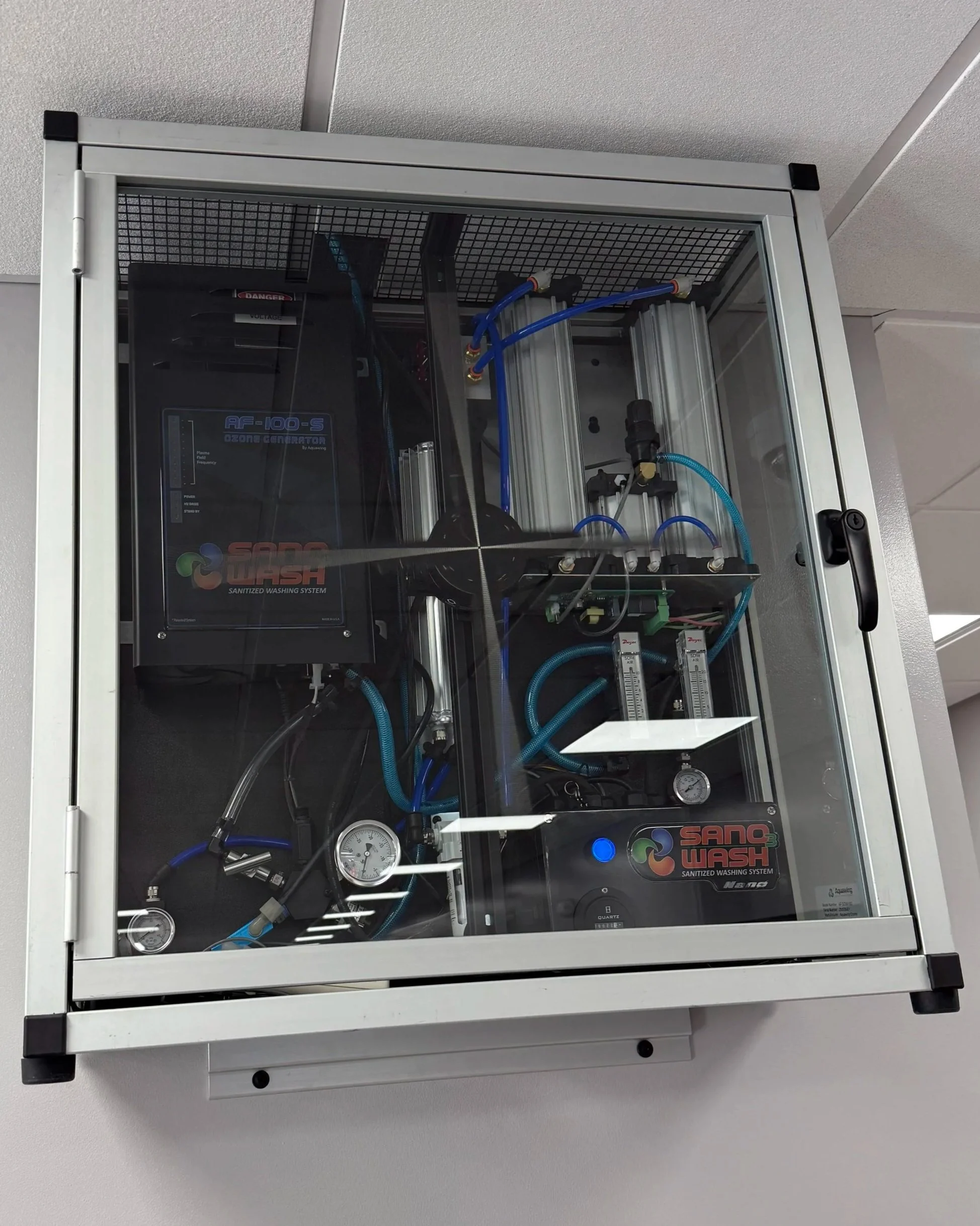 A sanitized water-based industrial washing system enclosed in a metal and glass cabinet with visible tubing, gauges, and electronic components.