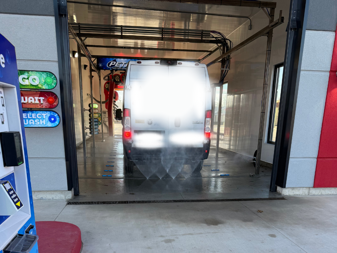 A white delivery van inside an automated car wash station with water spray on the rear end, and the wash equipment visible at the top.