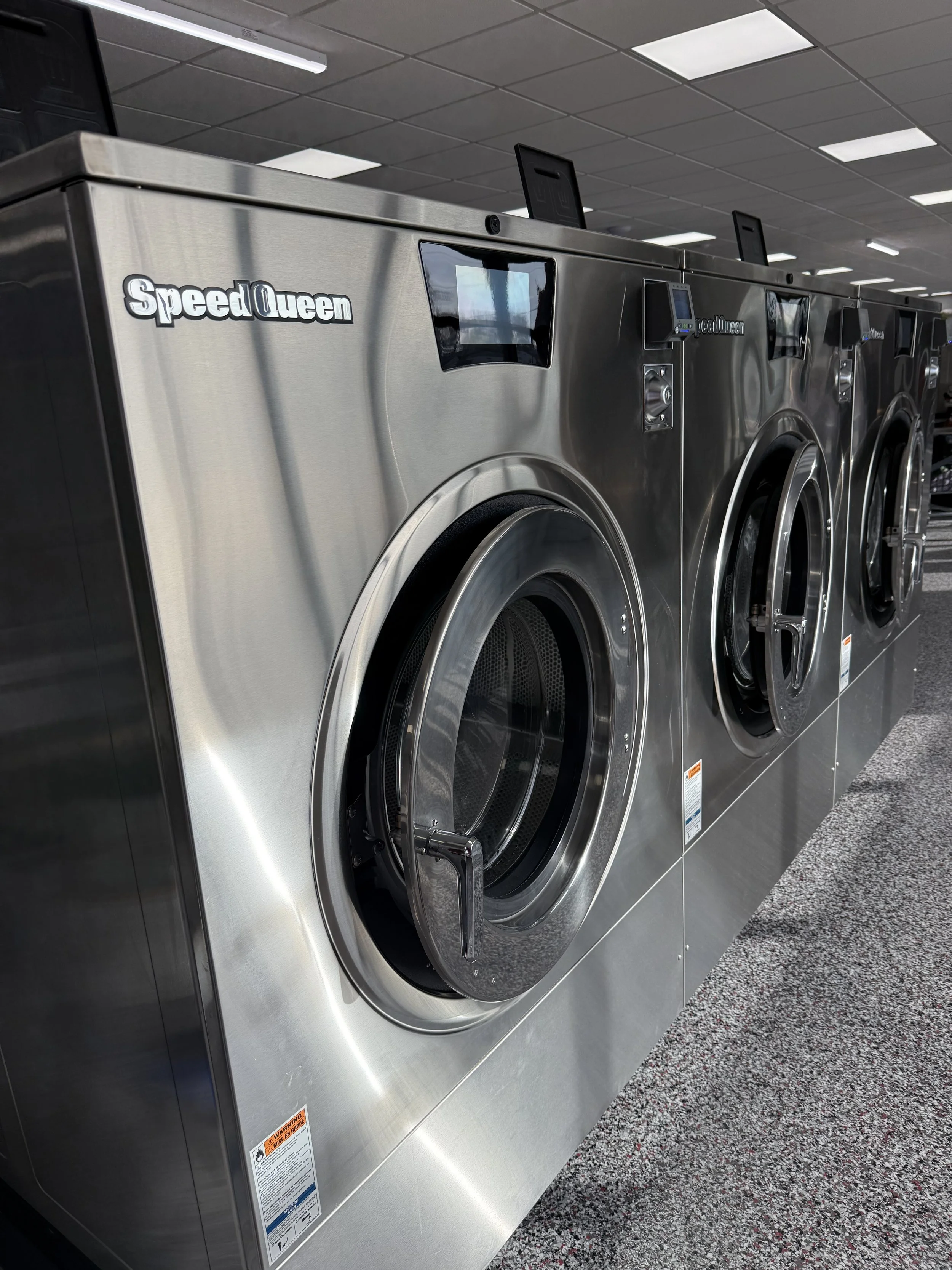 Row of commercial stainless steel Speed Queen washing machines in a laundromat, with digital controls and large round doors, set in a tiled room with ceiling lights.