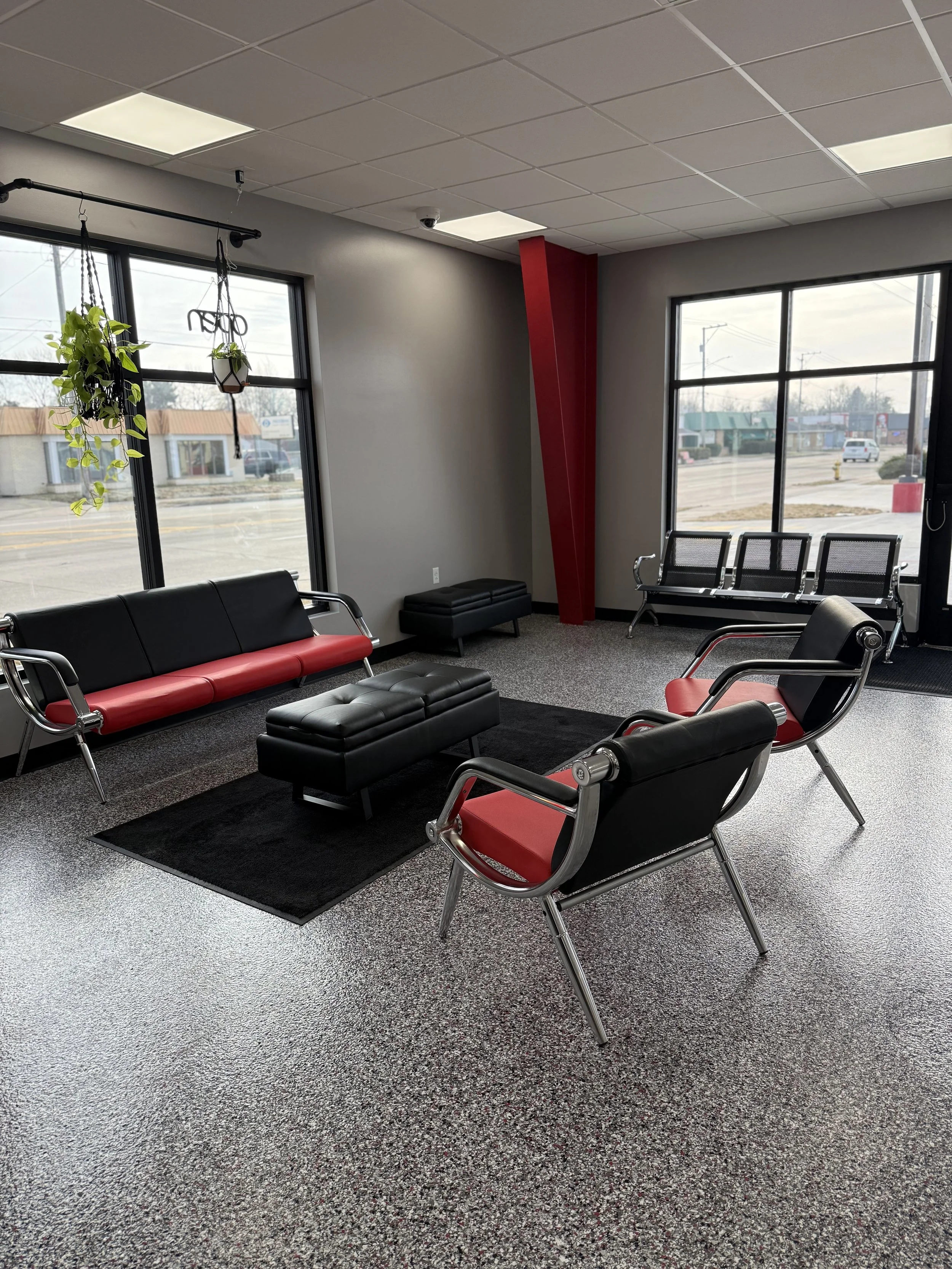 Modern waiting room with black and red chairs, a black leather ottoman, hanging plants, large windows, and a gray carpeted floor.