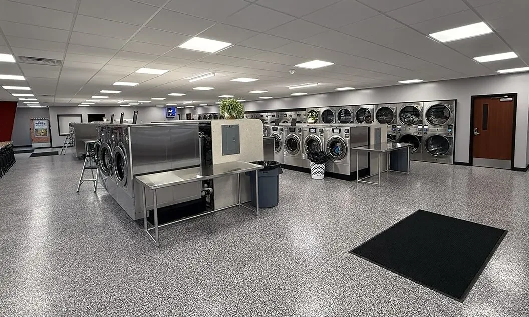 laundry room with multiple washing machines and dryers, stainless steel tables, a black mat, and a trash bin