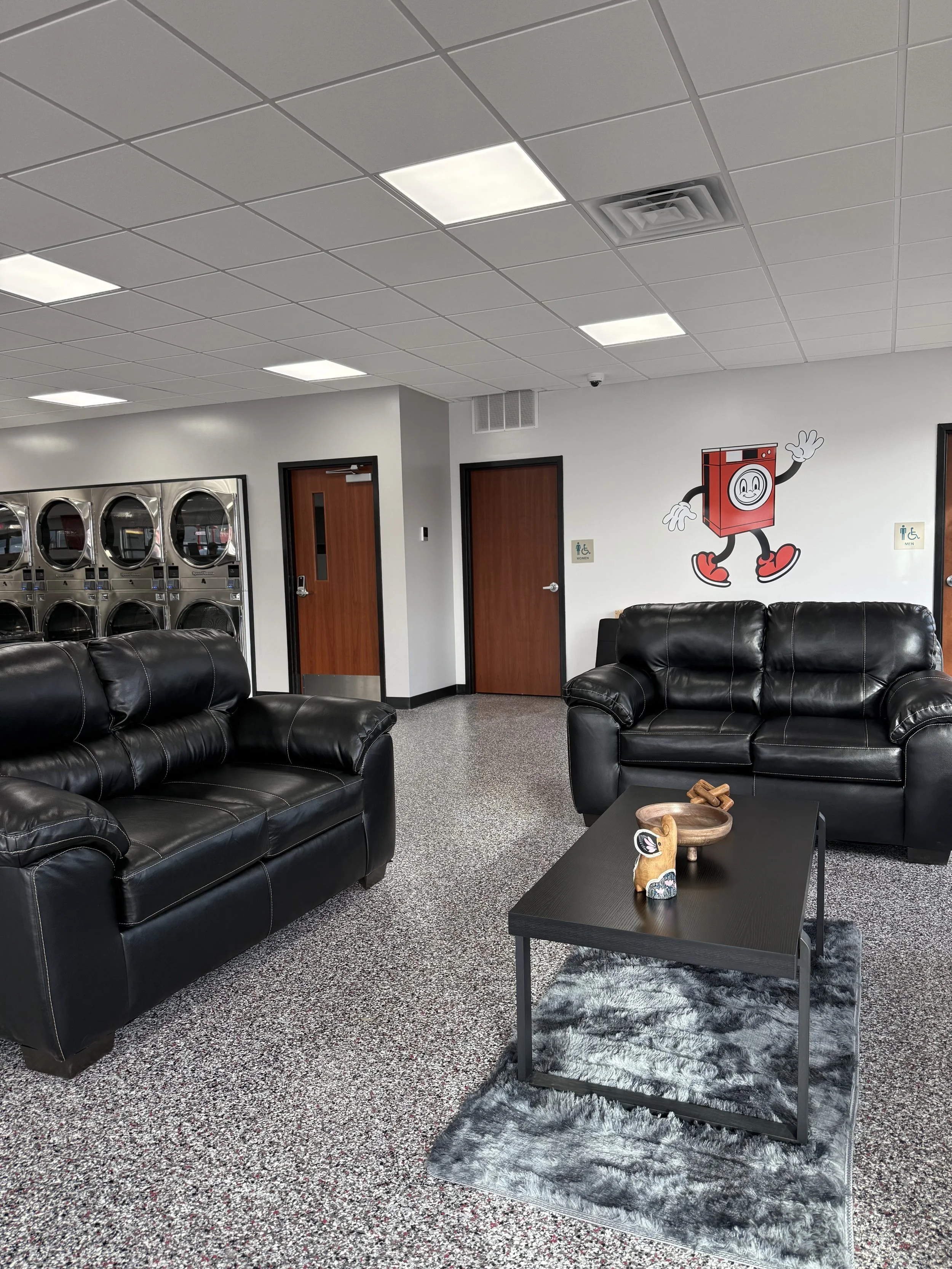 A waiting room with two black leather sofas, a black coffee table with decorative items, a gray furry rug, a wall with a cartoon washing machine character waving, a patterned carpet, stainless steel laundry machines, and wooden doors.