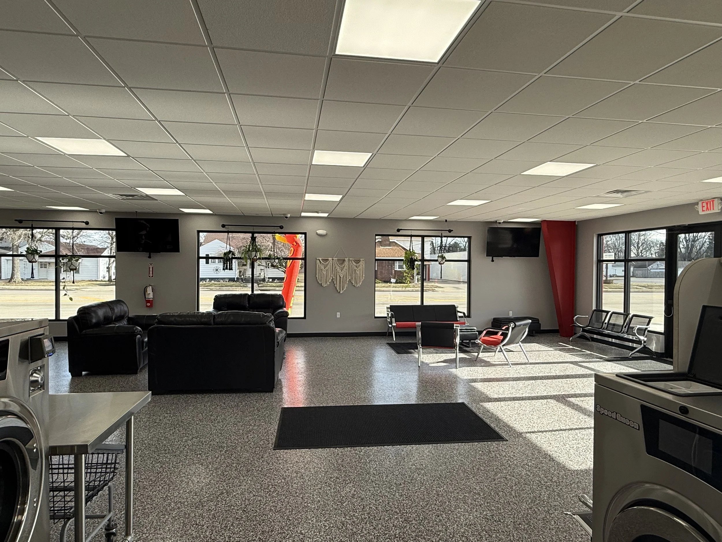 Interior of waiting room with black leather couches, benches, and chairs, large windows, potted plants, and wall-mounted TVs, in a commercial building.