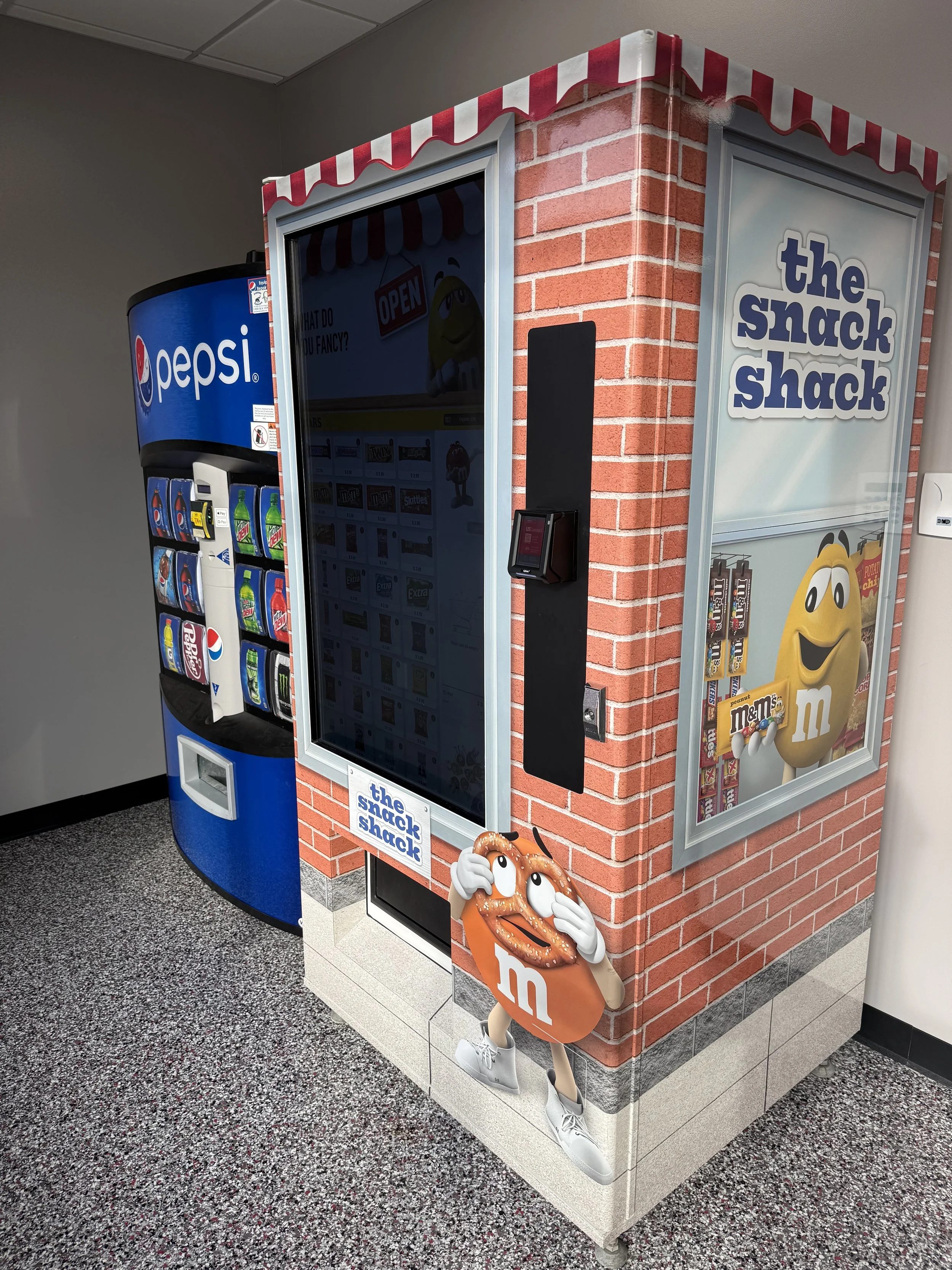 A corner kiosk designed to look like a small snack shop with a brick exterior, branded as 'the snack shack.' It features a large touchscreen, a nearby vending machine for Pepsi and other drinks, and cartoon decorations, including an M&M character and a pretzel character with white sneakers.