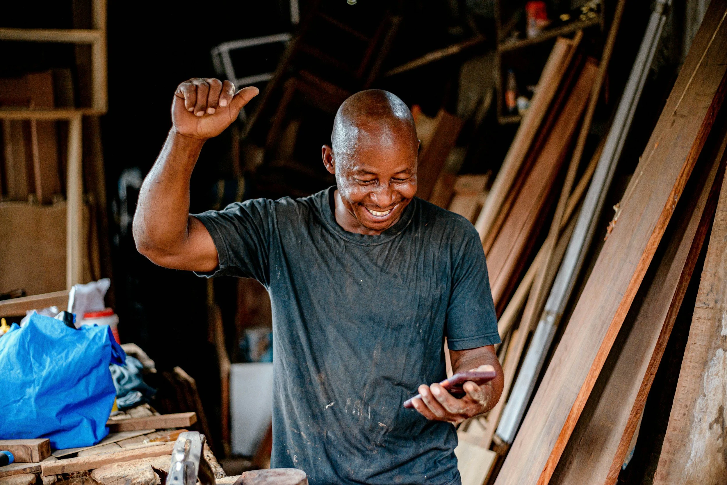 A man smiling and raising one hand in a woodworking shop while looking at his phone.