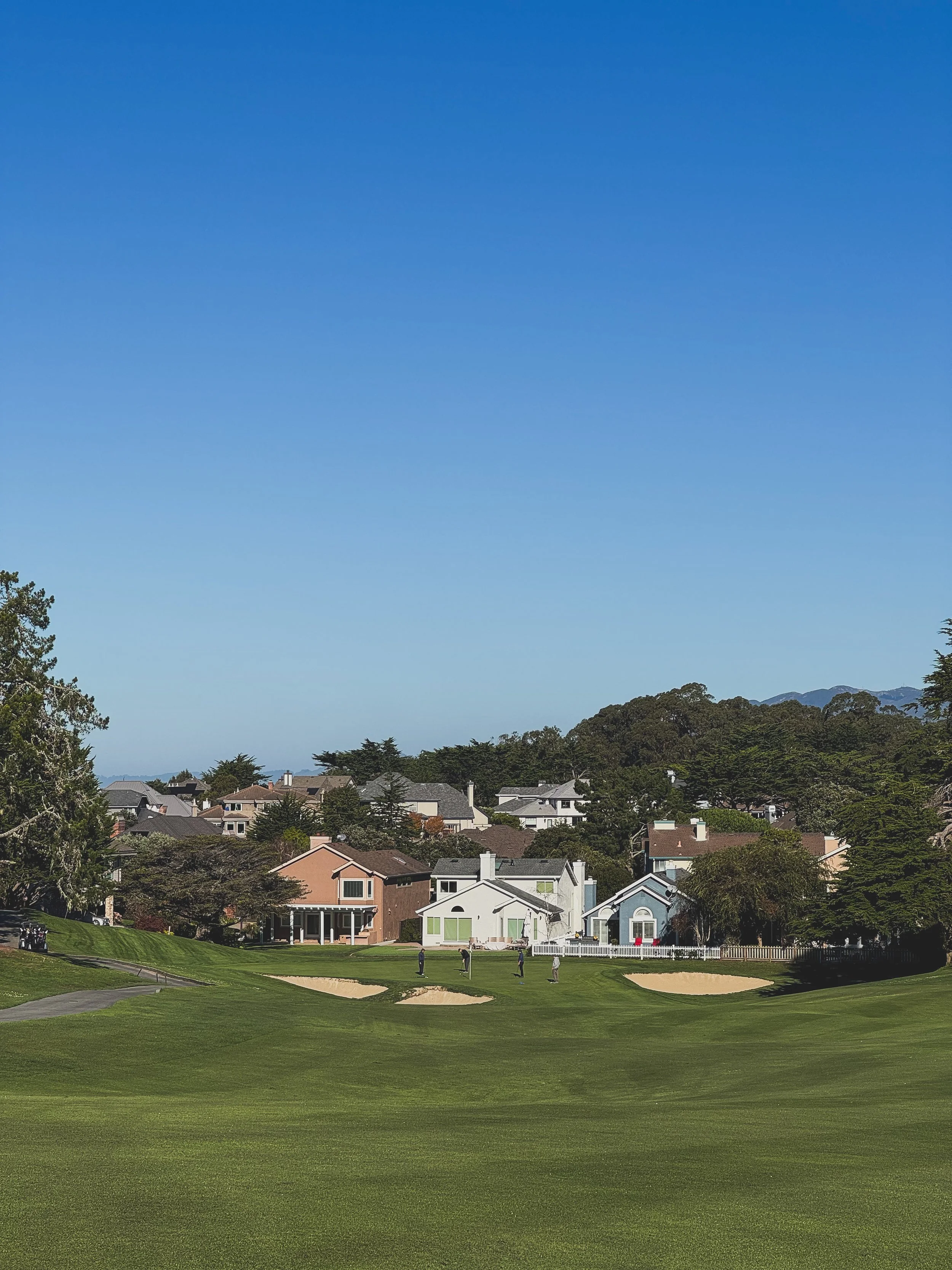 A golf course with a few people playing, lush green grass, sand bunkers, and houses in the background under a clear blue sky.