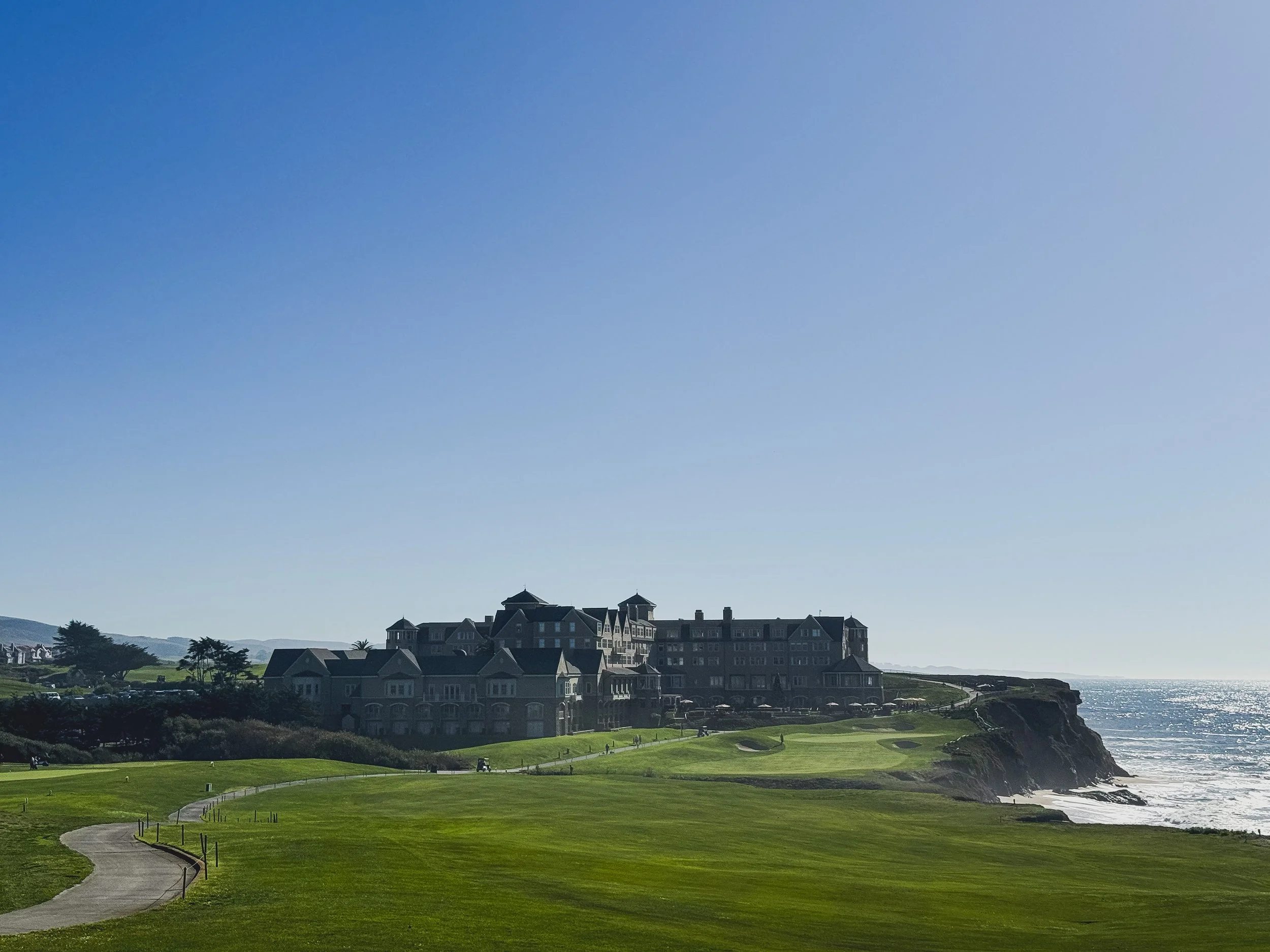 A large beachside hotel surrounded by a lush green golf course, with the ocean on the right and a clear blue sky above.