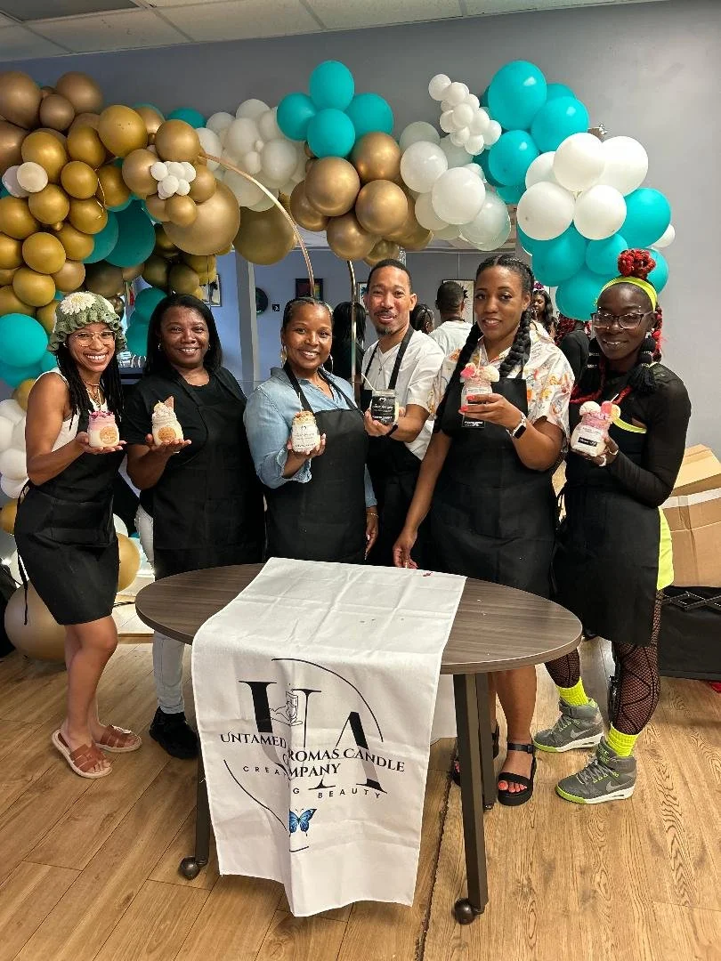 Six women standing behind a table holding candles, with colorful balloon arch in the background, at a celebration event for Untamed Aroma Candle Company.