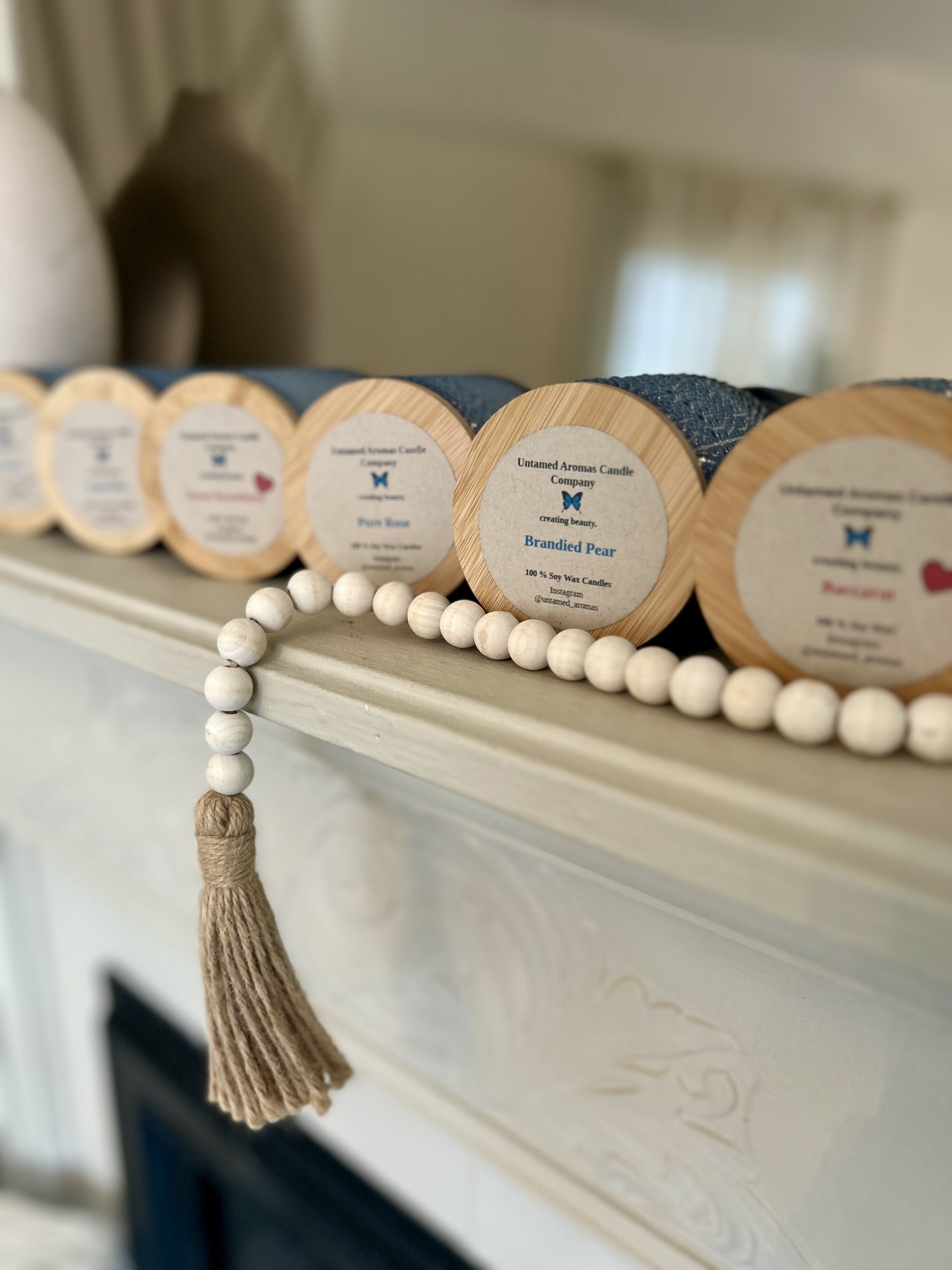 Close-up of candles labeled 'Untamed Aromas Candle Company,' with a focus on a candle named 'Brandied Pear,' a tassel with white beads hanging from the shelf.