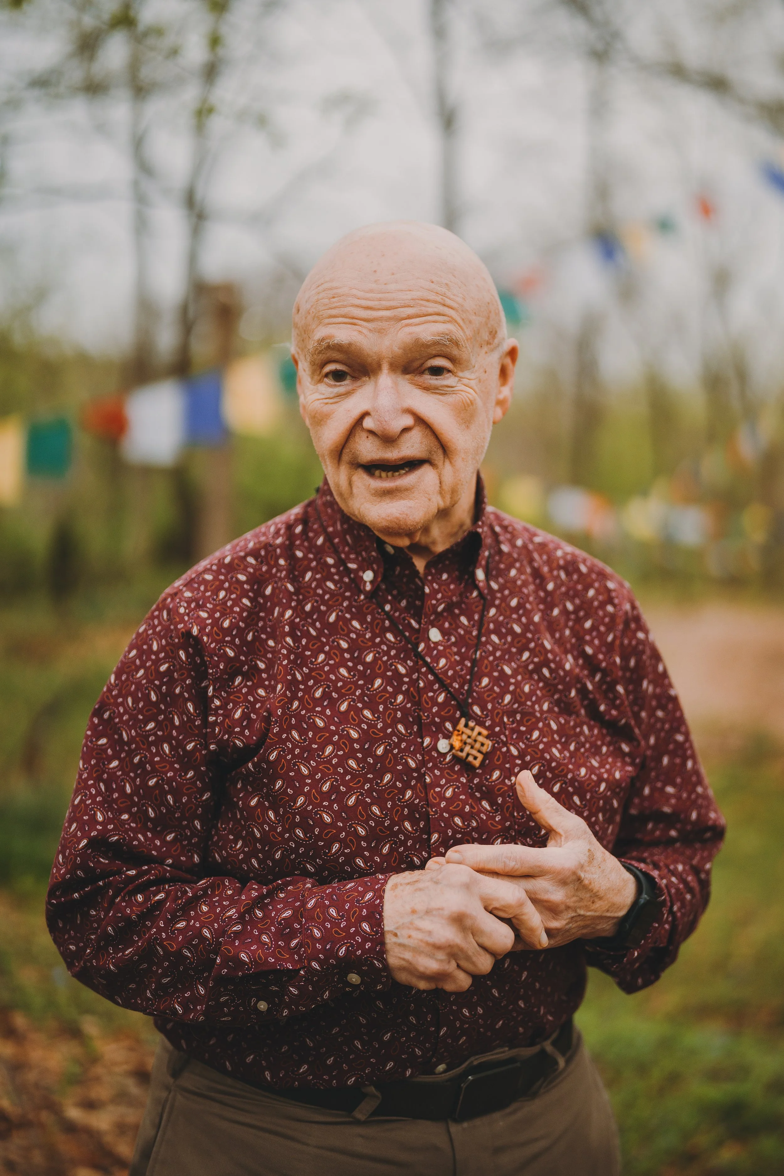 An elderly man with a bald head wearing a patterned red shirt and a necklace with a pendant, outdoors near trees with colorful flags hanging in the background.