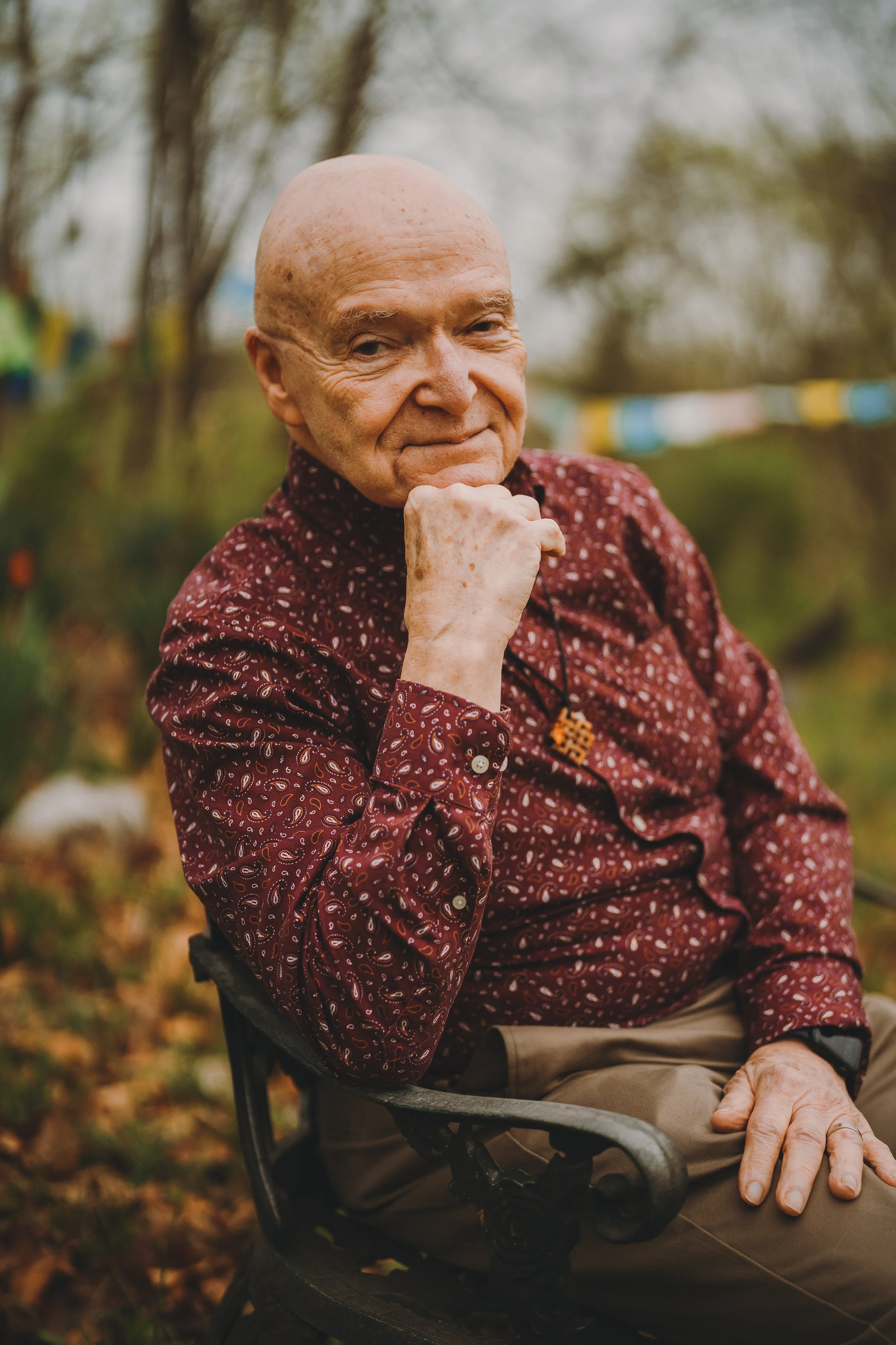 An elderly man sitting outdoors, wearing a patterned maroon shirt, with a hand resting on his chin and a gentle smile.