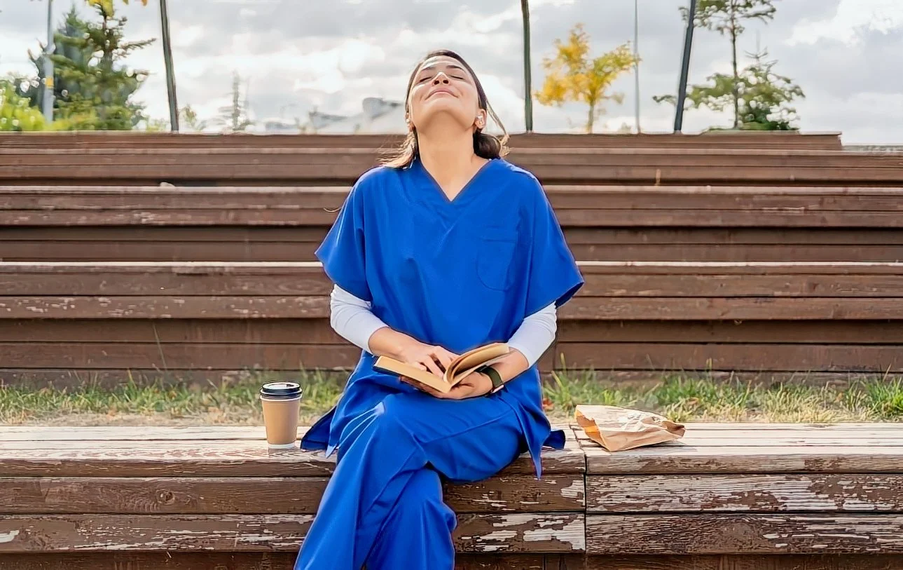 A woman sitting outdoors on a wooden bench, wearing a blue medical uniform, with her eyes closed and head tilted back, holding an open book in her lap. There is a coffee cup on her right and a paper bag on her left.