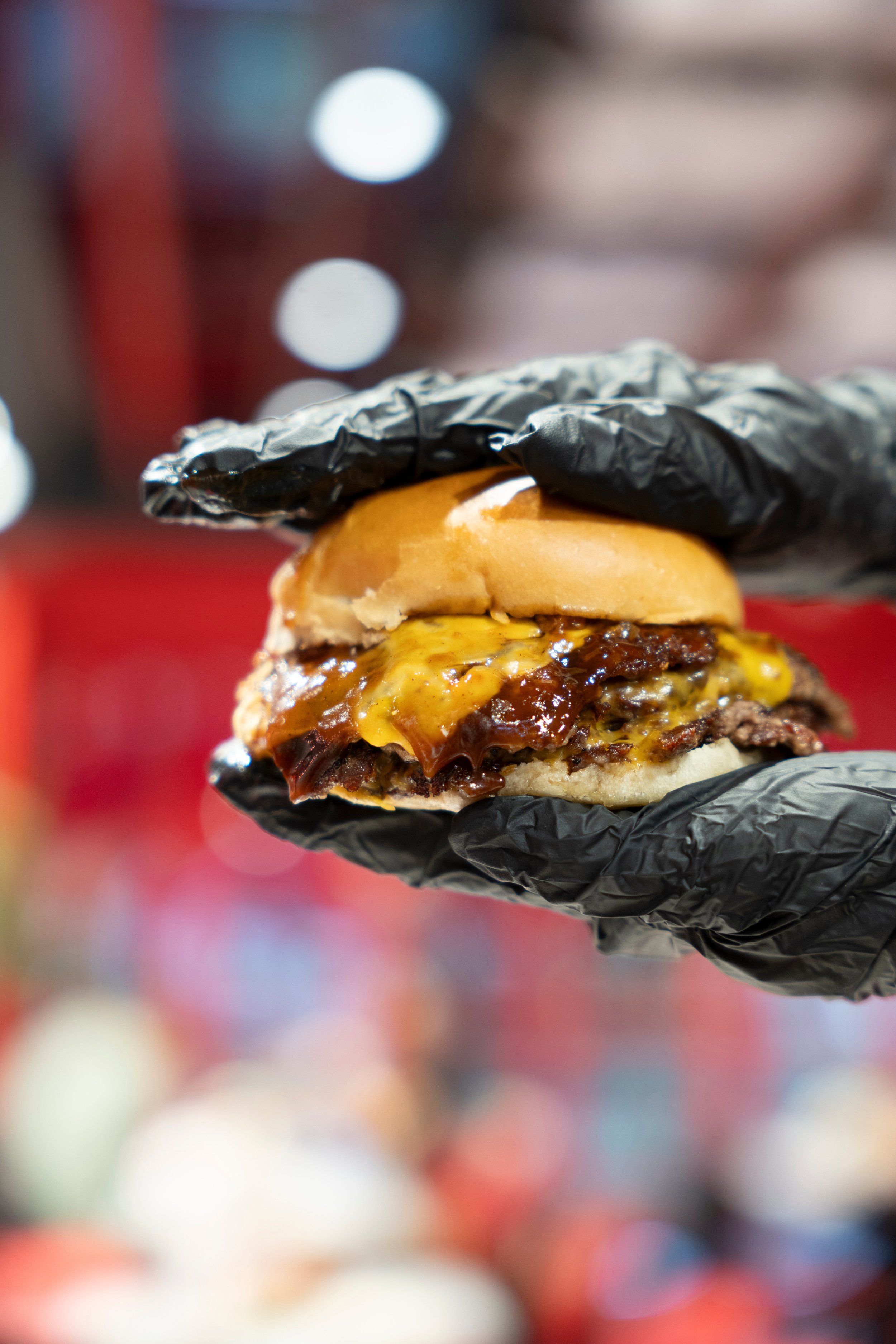A person wearing black gloves holding a cheeseburger with lettuce, cheese, and barbecue sauce in a bun.