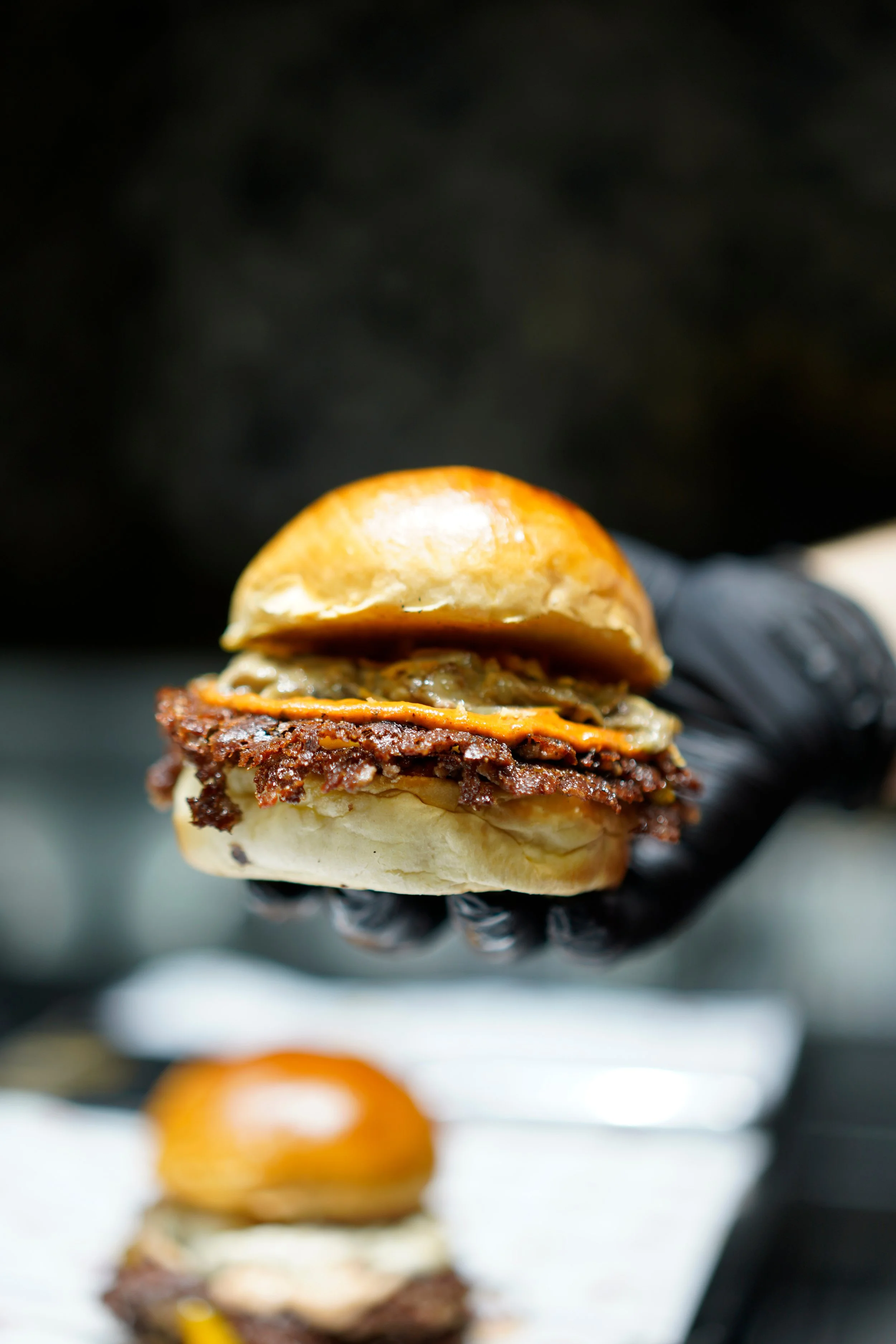 A close-up of a burger being held in a hand with a black glove. The burger has a bun, a beef patty, melted cheese, and crispy bacon.