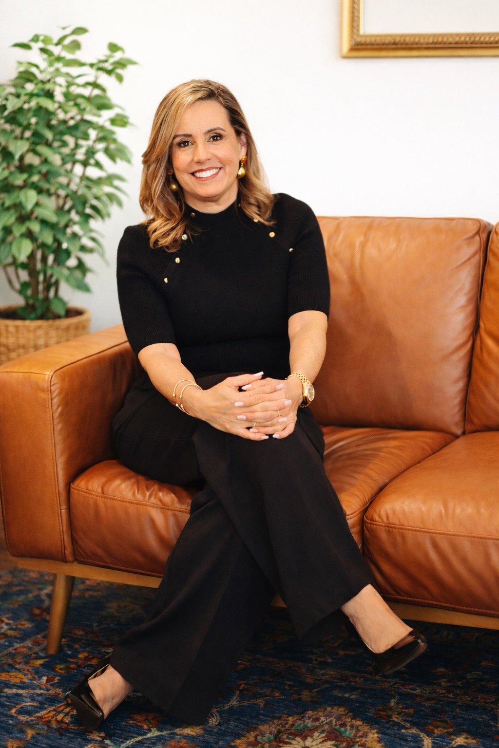 A woman with shoulder-length light brown hair sitting on a tan leather couch. She is smiling, wearing a black top with gold buttons, gold earrings, a gold watch, and black pants. There is a green potted plant behind her and a framed picture on the white wall.