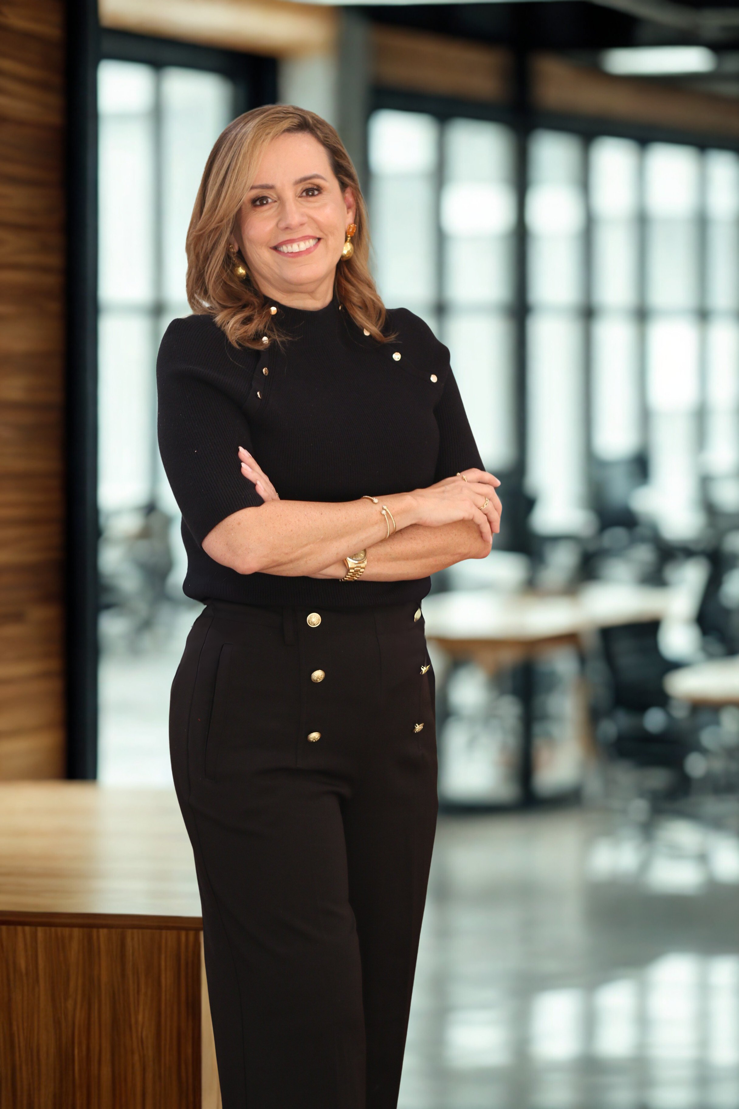 A woman with shoulder-length brown hair standing with her arms crossed, wearing a black top and black pants with gold buttons in a modern office setting.