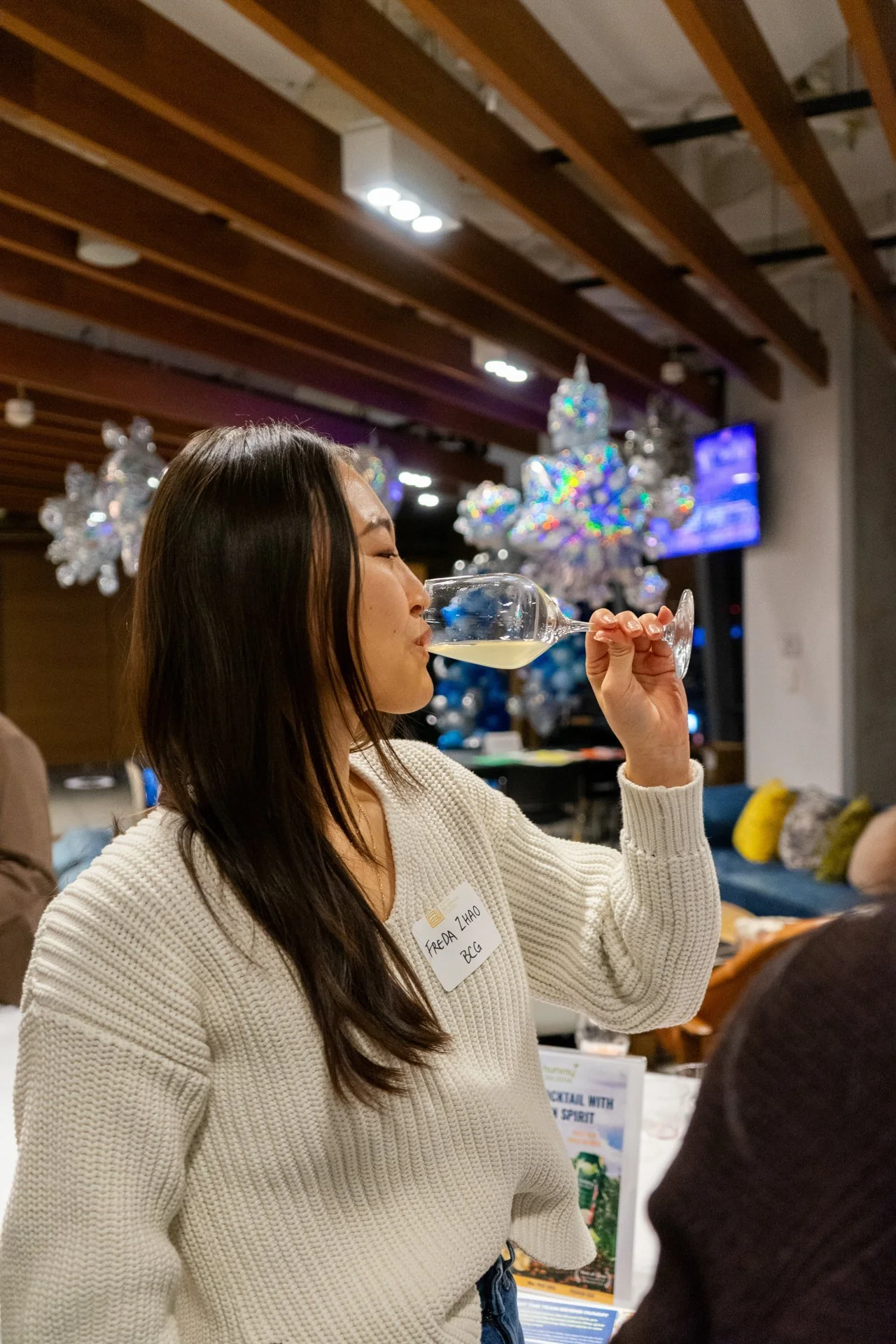 A woman with long dark hair wearing a white sweater is holding a wine glass and taking a sip. In the background, there are holiday decorations including snowflake ornaments and a decorated Christmas tree.