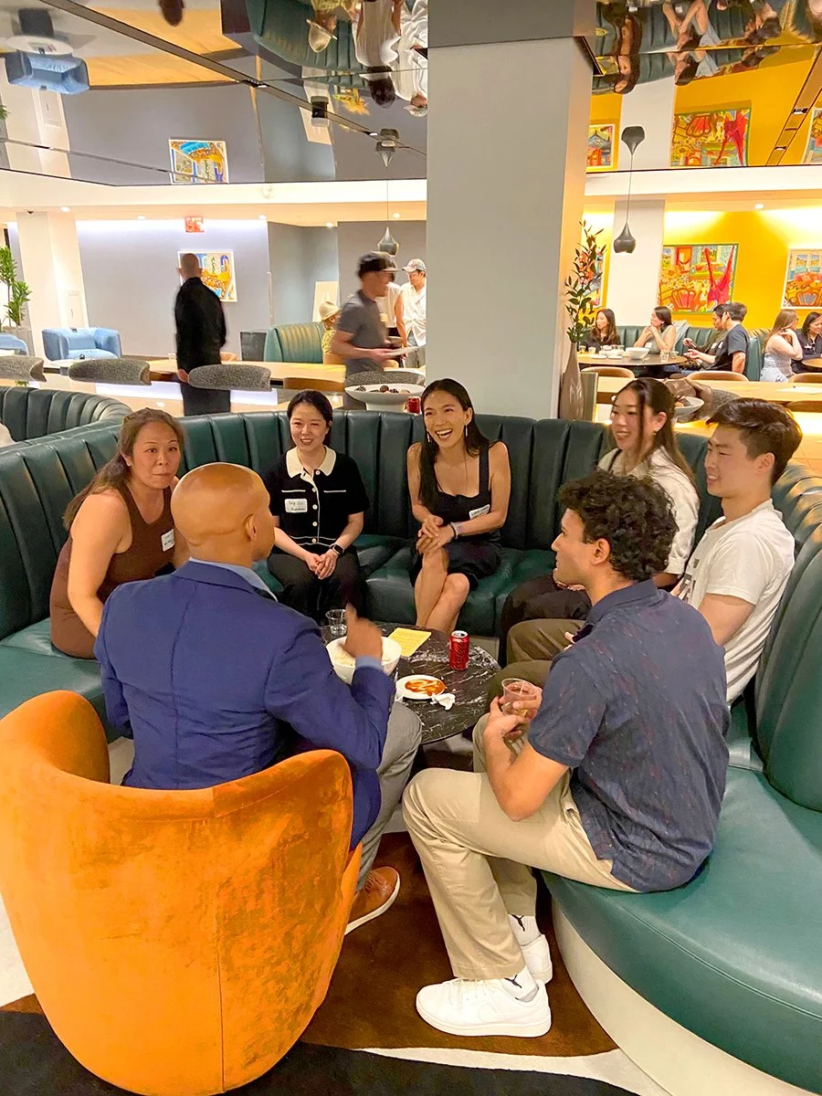 Group of people sitting in a semi-circle on a couch and chairs in a restaurant or cafe, engaged in conversation, with others dining in the background.