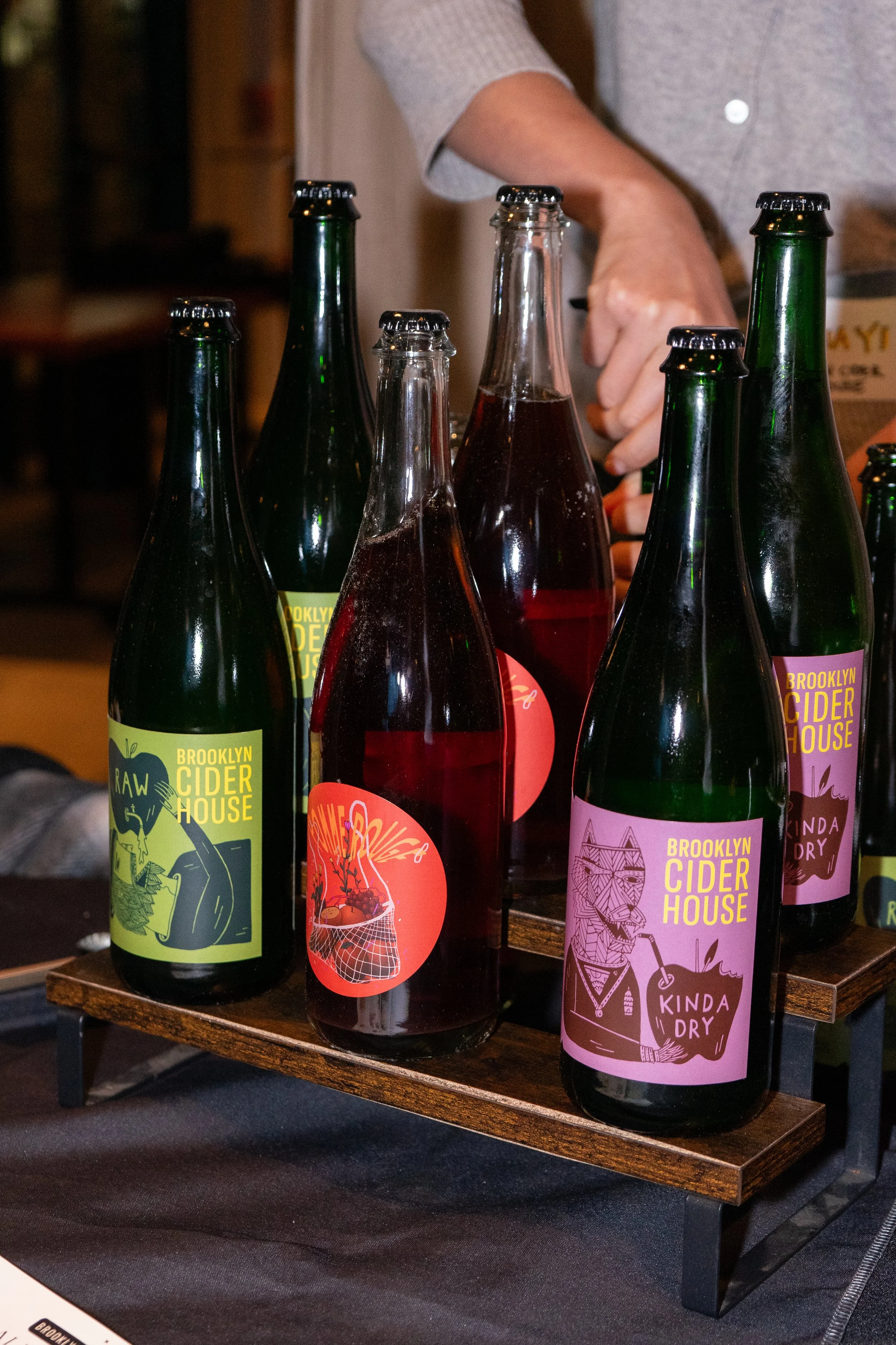 Assorted bottles of Brooklyn Cider House on a wooden tray, with colorful labels featuring graphic designs, on a black table.